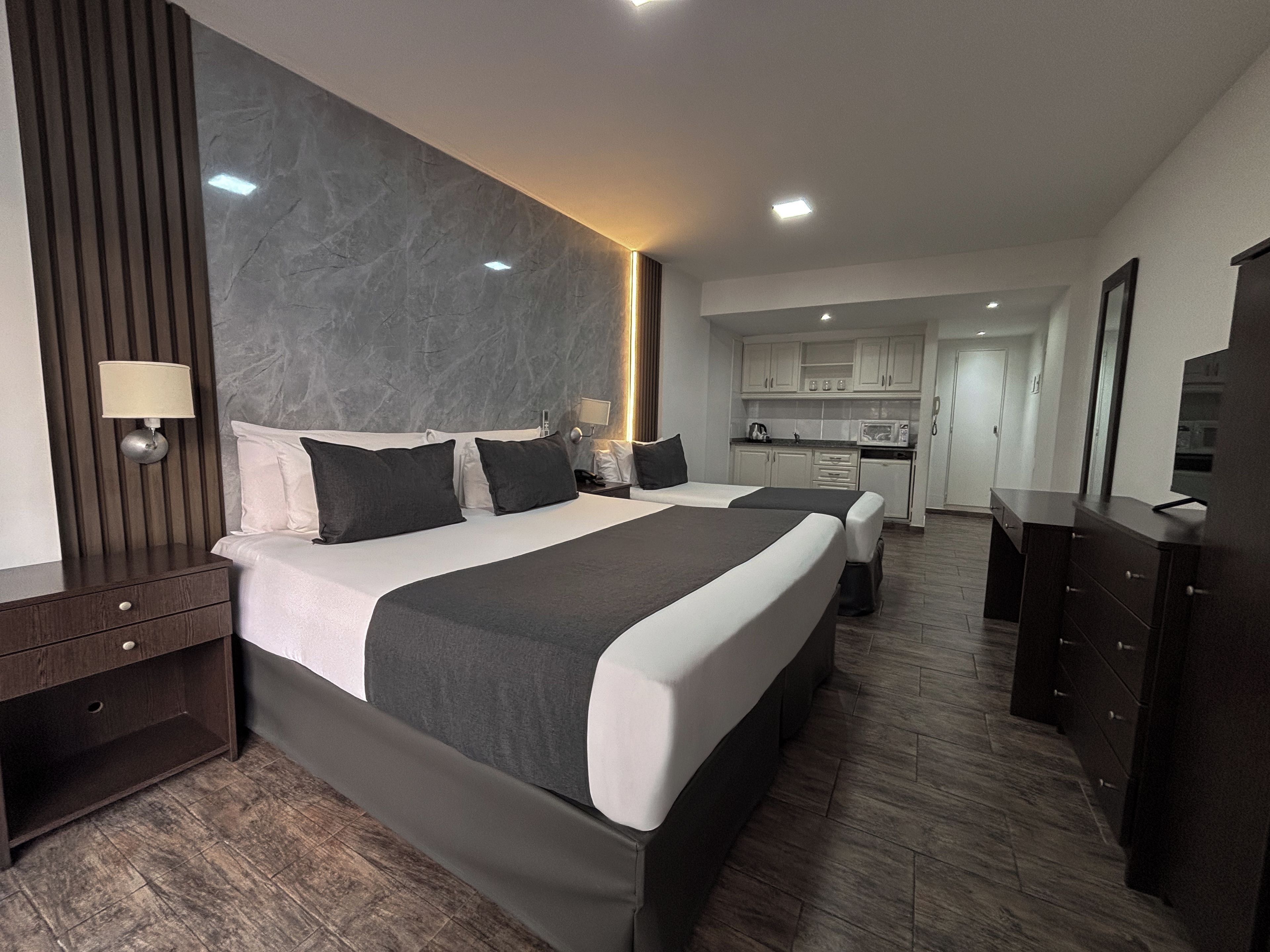 A modern hotel room with a large bed, a bedside table, a wardrobe, and a kitchenette in the background.