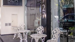 The exterior of the KER Belgrano Hotel with a glass facade, white metal chairs, and a signboard.