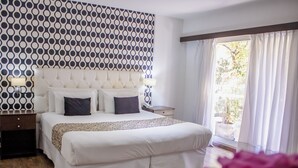 A hotel room with a large bed, a patterned wall, a nightstand, and a view of the outdoors.
