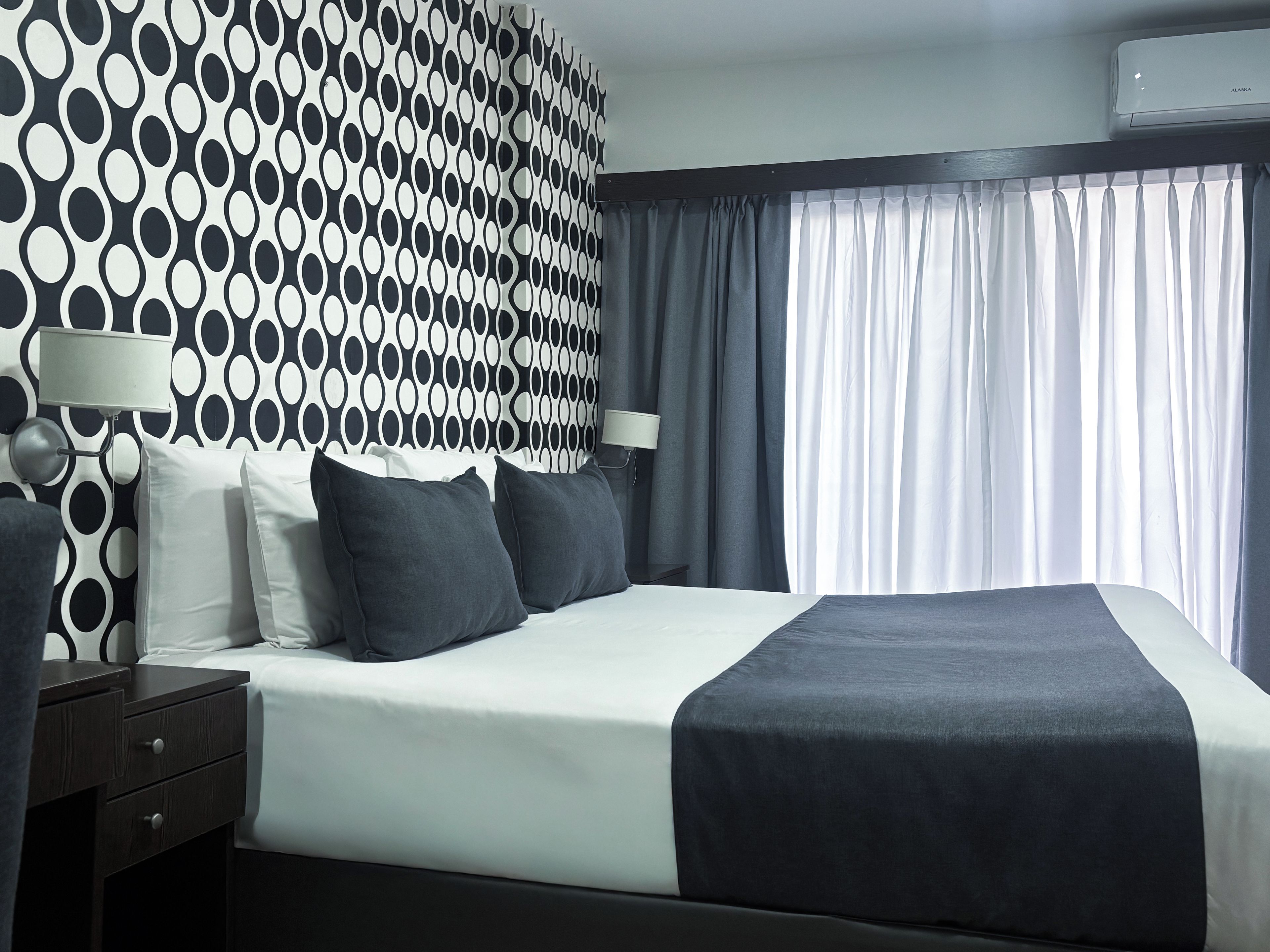 Habitación de hotel con una cama grande, dos mesitas de noche, una pared con un diseño circular blanco y negro, y una ventana con cortinas.
