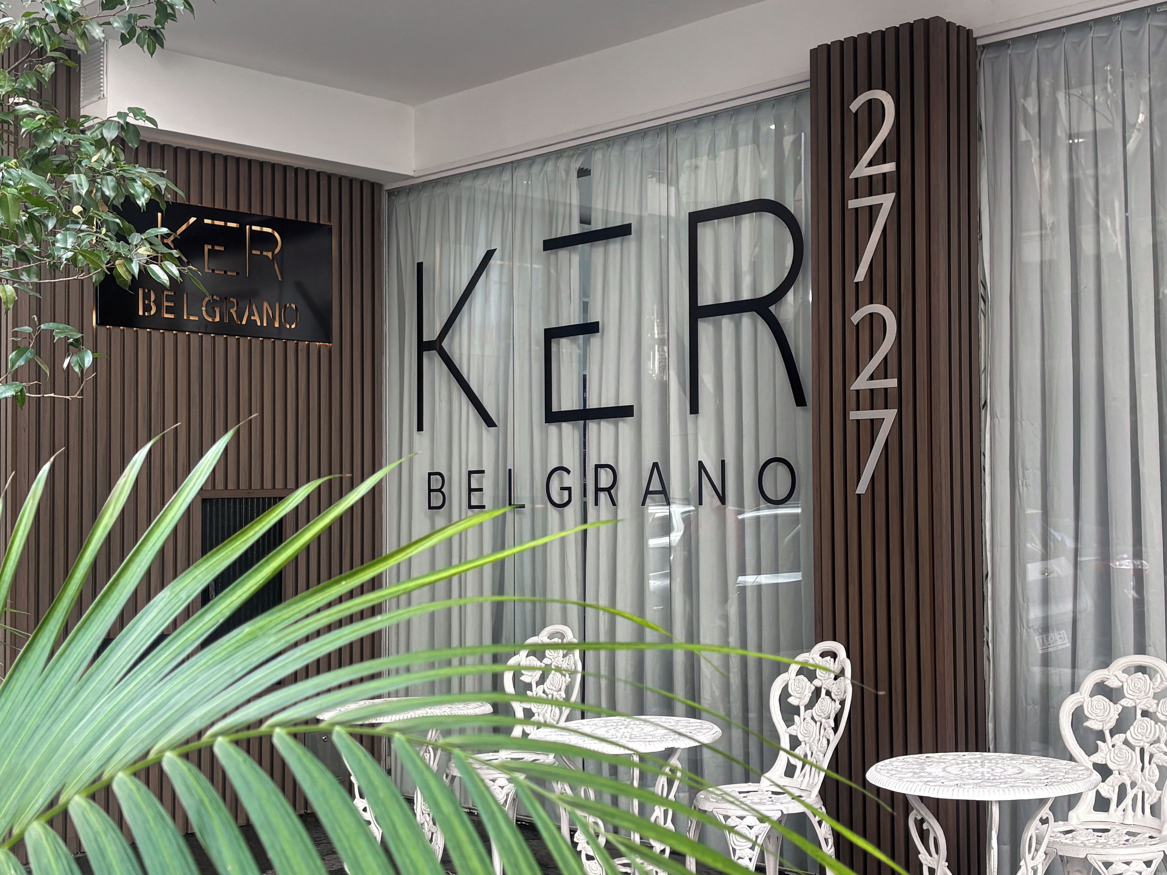 Interior moderno com uma placa grande indicando 'KER BELGRANO' e o número '2727'. Cadeiras brancas e uma mesa pequena em primeiro plano.