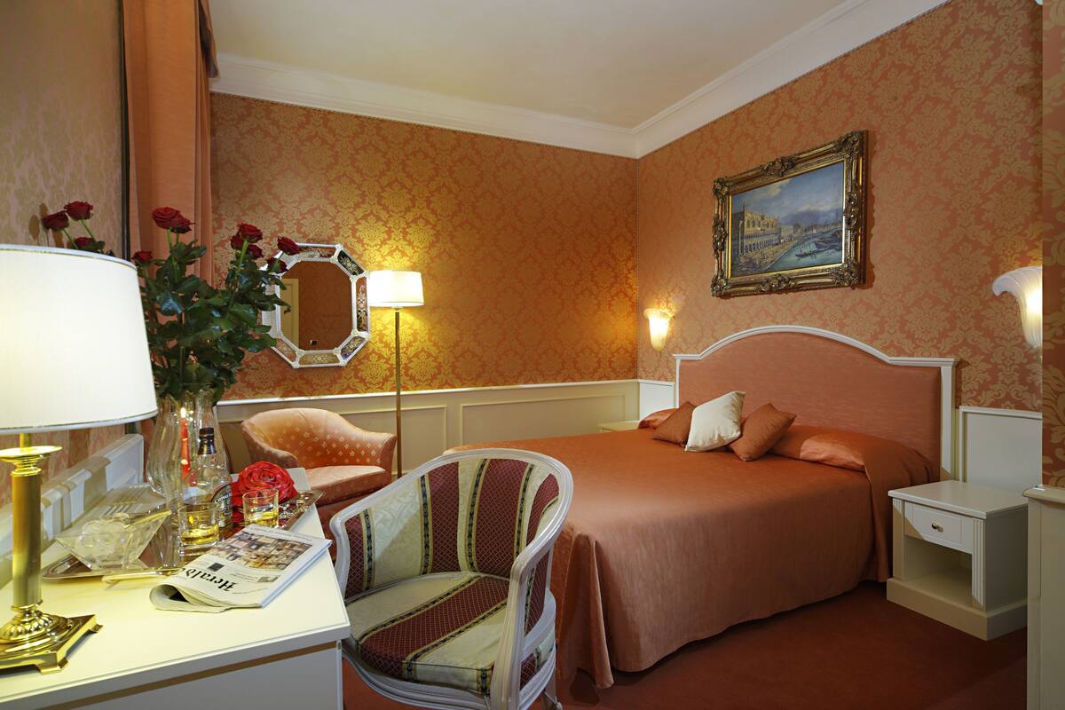 Ein Hotelzimmer mit einem Bett, einem Sessel, einem Schreibtisch mit Lampe, einer Vase mit Blumen und einem Bild an der Wand.