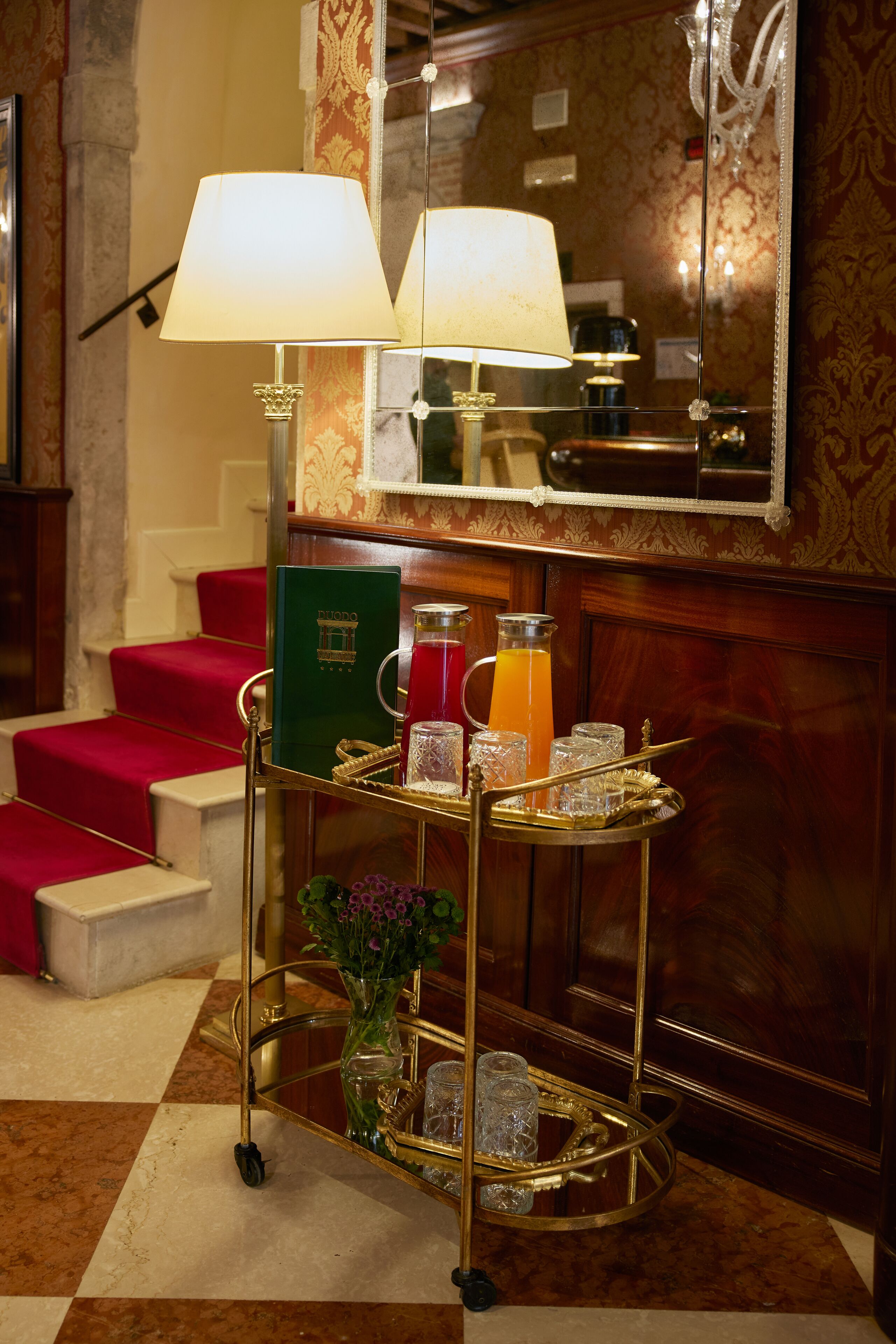 Vestíbulo de hotel con una escalera alfombrada de color rojo, un espejo con marco dorado y un carrito de bar con bebidas y un libro verde.