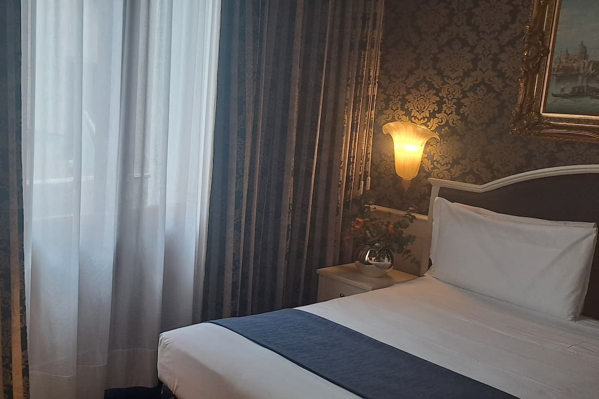 Ein Hotelzimmer mit Bett, Vorhängen, einem Nachttisch, einer Lampe und einer Wandleuchte.