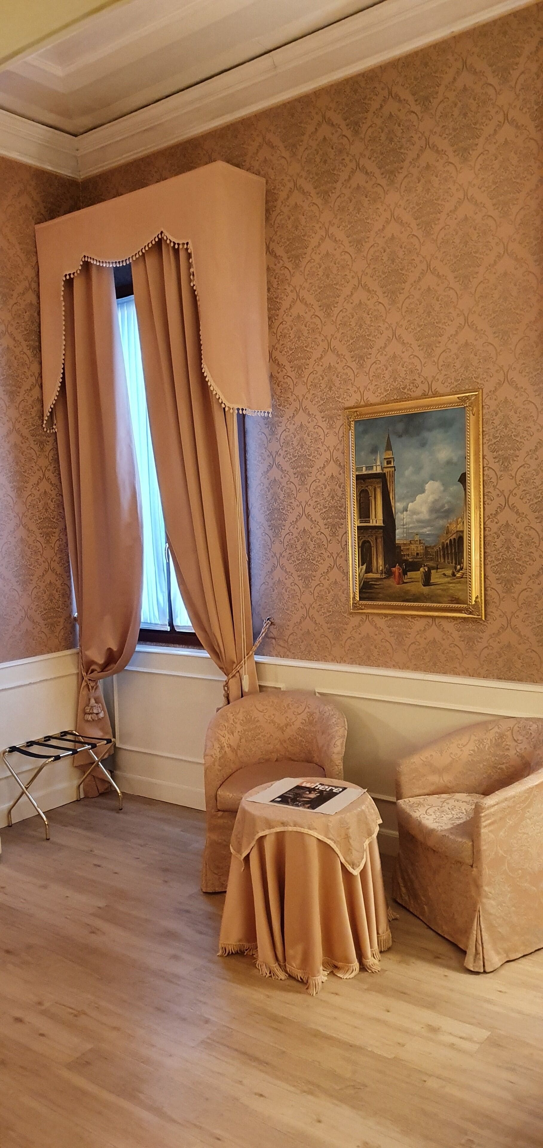 A room with patterned wallpaper, a window with curtains, a framed painting, and a small table with a magazine.