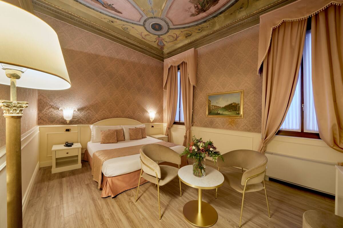 Ein Hotelzimmer mit Bett, Nachttisch, Lampe, Stuhl, rundem Tisch und einer Blumenvase.