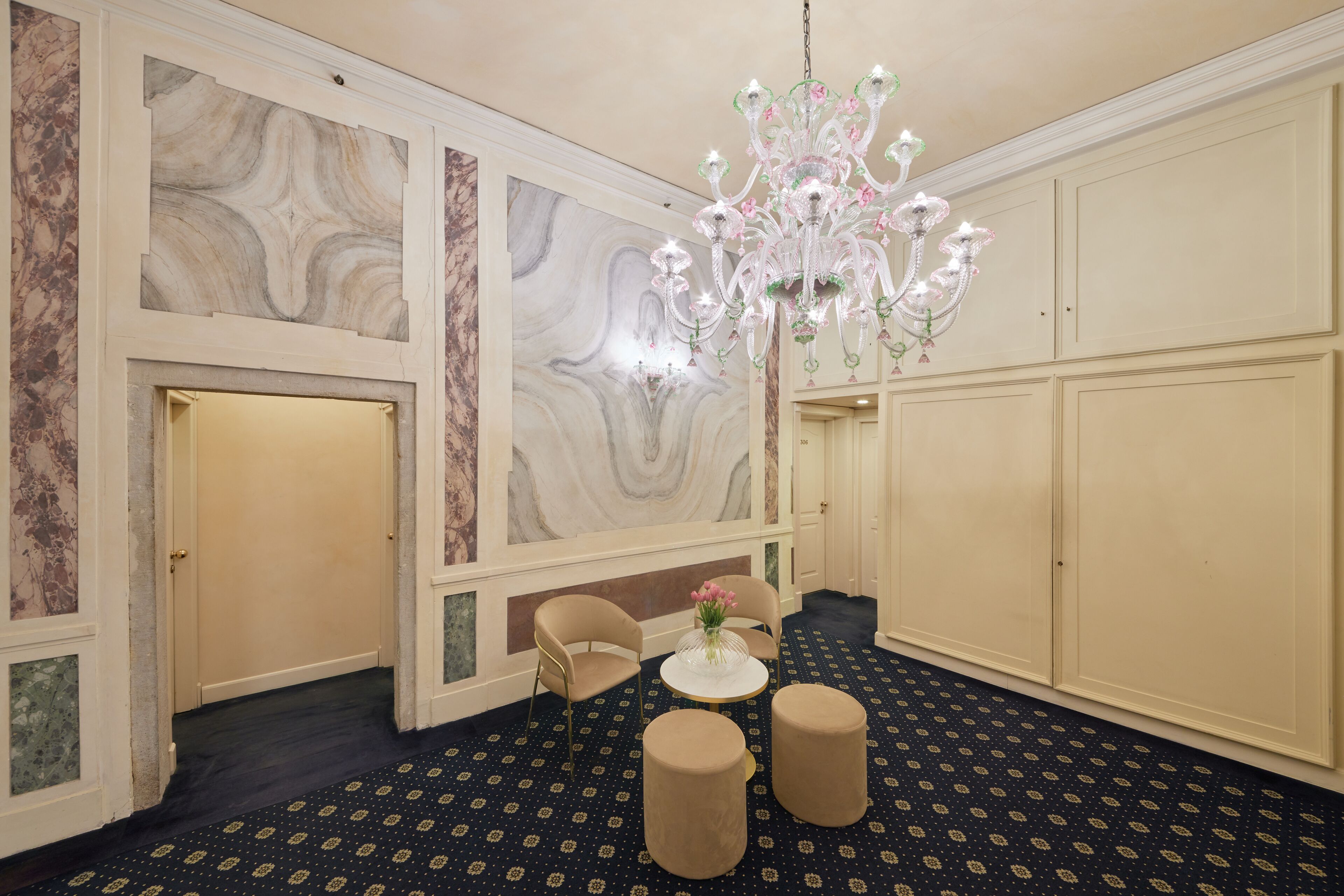 A room with a marble wall, a chandelier, two chairs, a small table, and a patterned carpet.