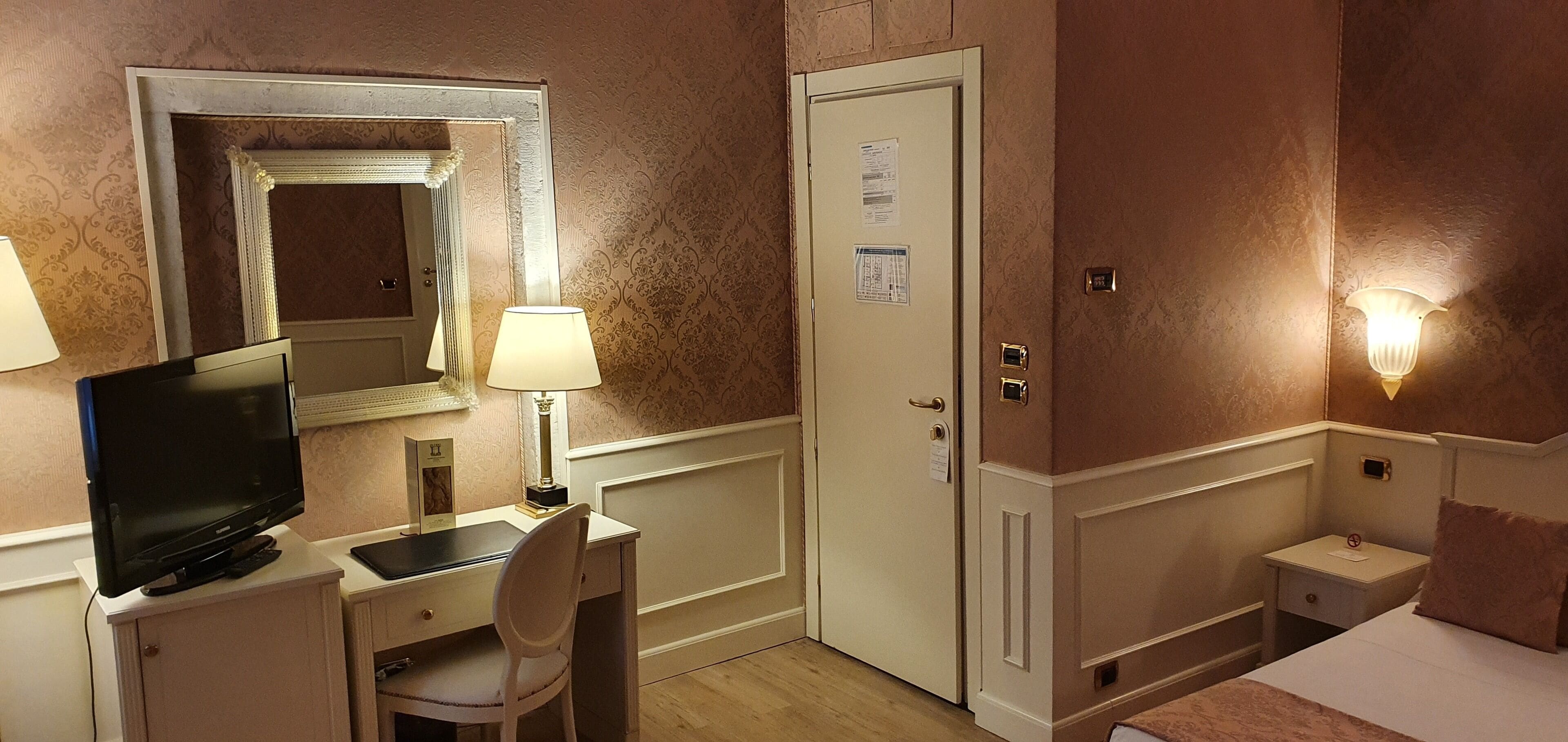 Habitación de hotel con escritorio, silla, televisión, lámpara, cama y puerta con un espejo encima.