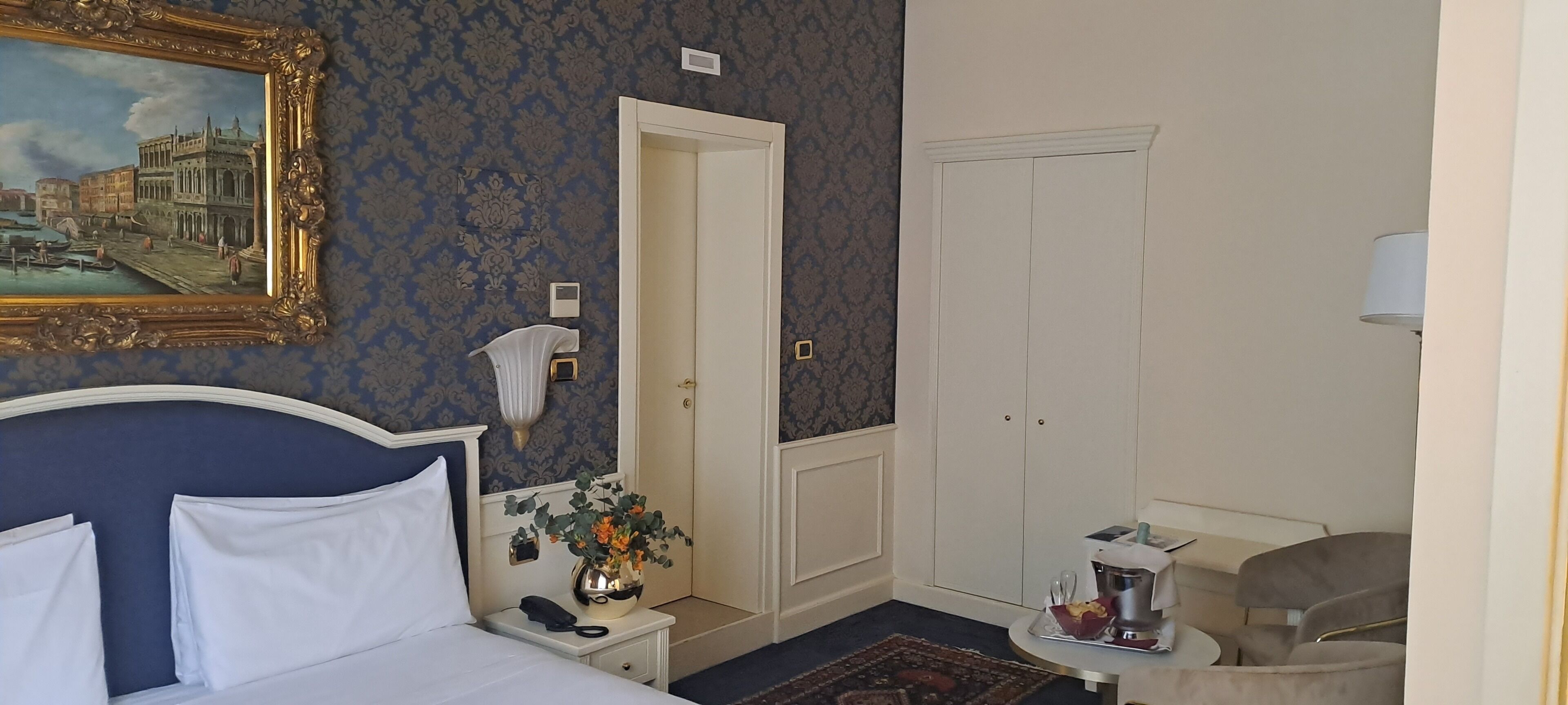 A hotel room with a bed, a nightstand with a vase of flowers, a desk with a lamp, and a door.
