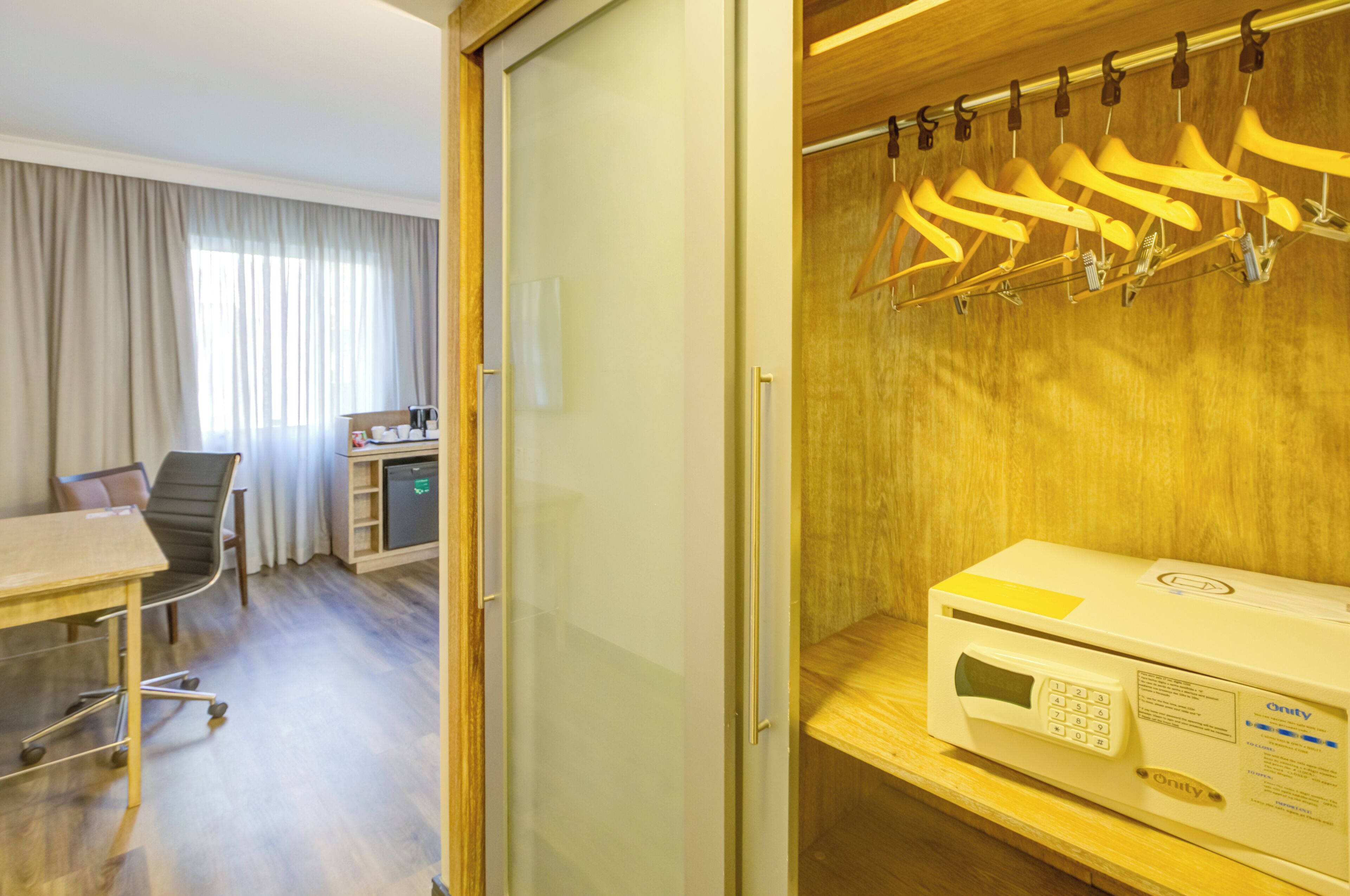 A hotel room with a wooden floor, a desk with a chair, a bed, and a coat rack with hangers.