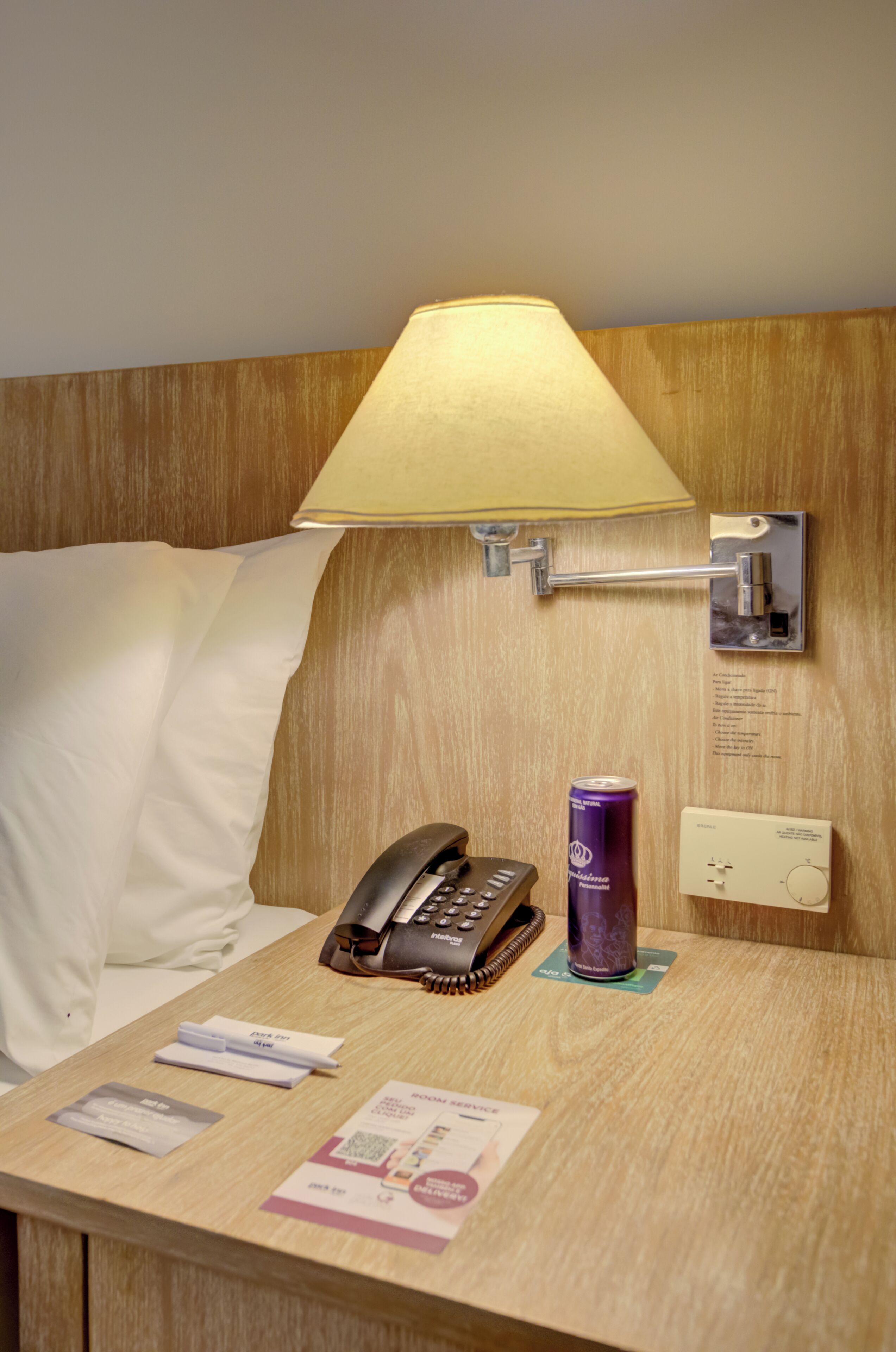 A hotel room with a bed, a bedside lamp, a telephone, a can of beverage, and a wall-mounted light switch.