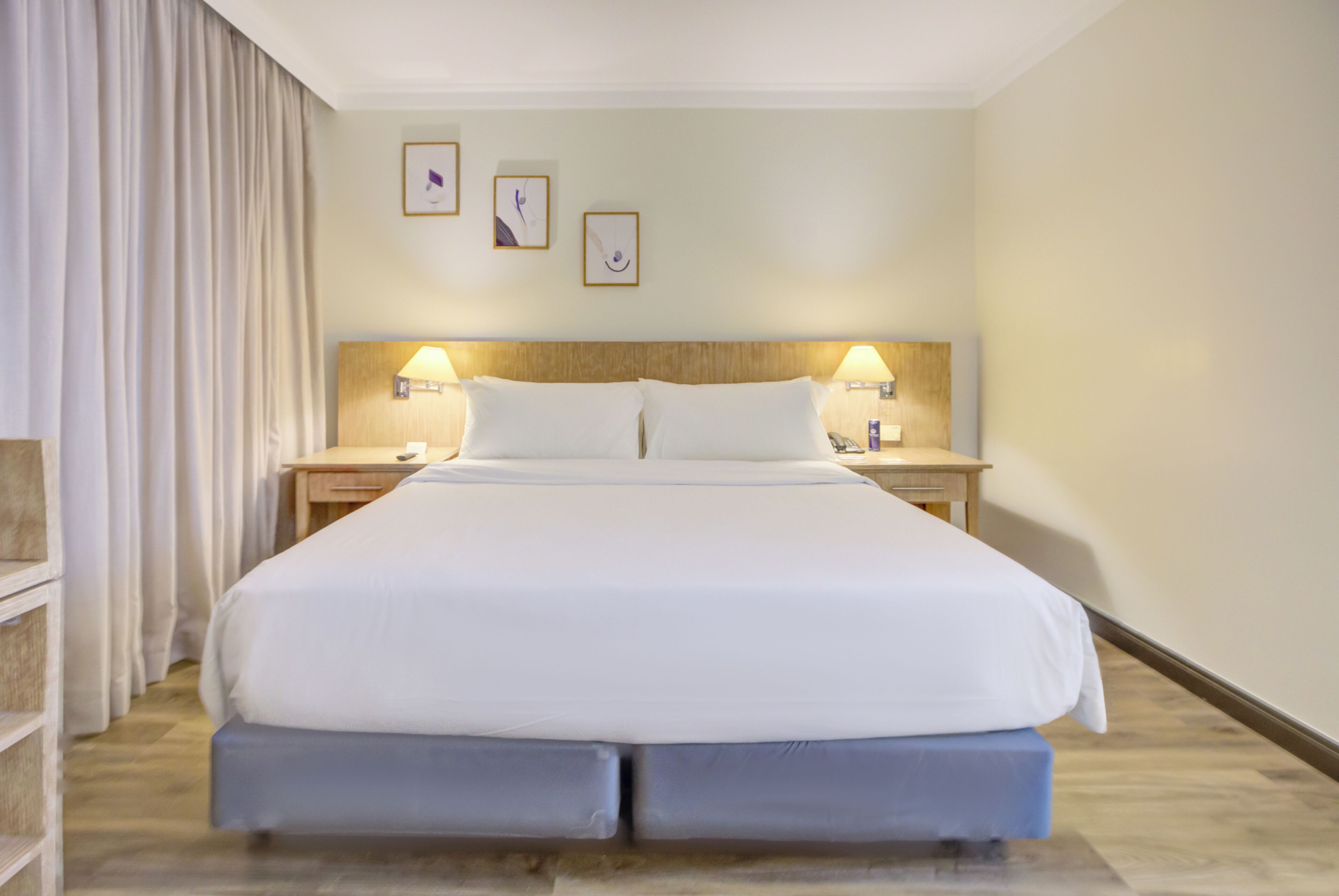 Hotellihuone, jossa on suuri sänky, kaksi yöpöytää valaisimineen, puinen pääty ja seinälle kiinnitetty taideteos.