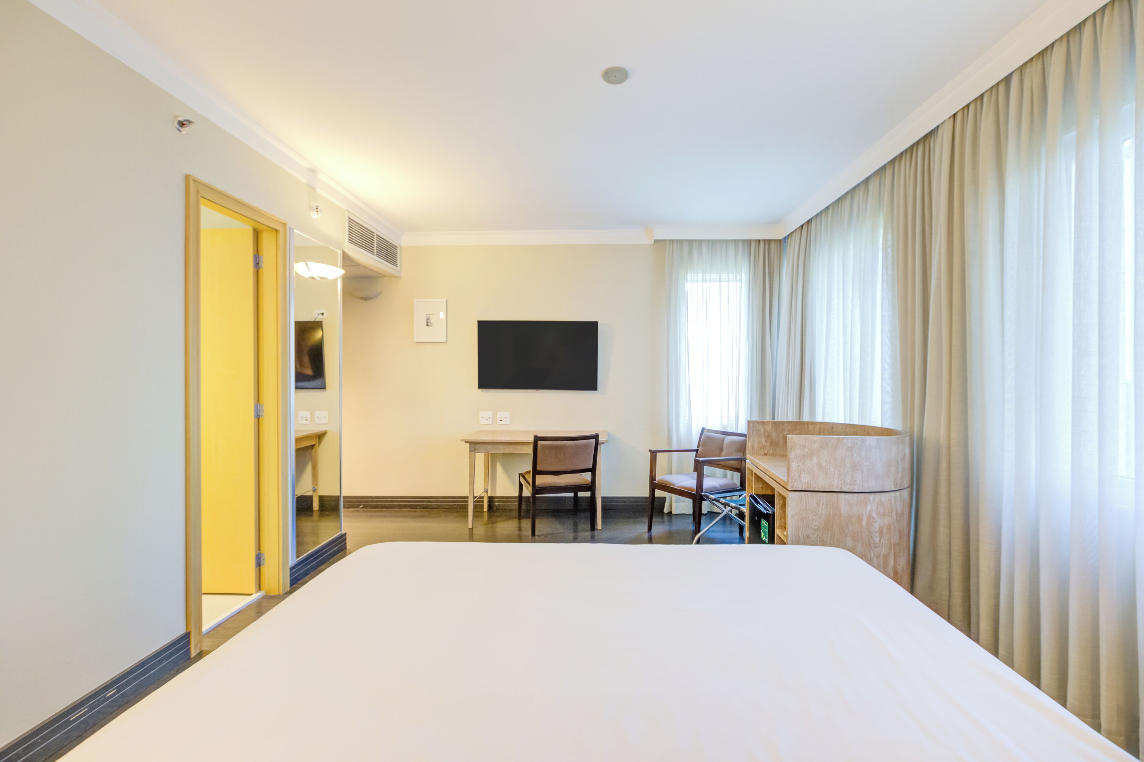 Habitación de hotel con una cama grande, un escritorio con dos sillas, un televisor de pantalla plana y un ventanal con cortinas.