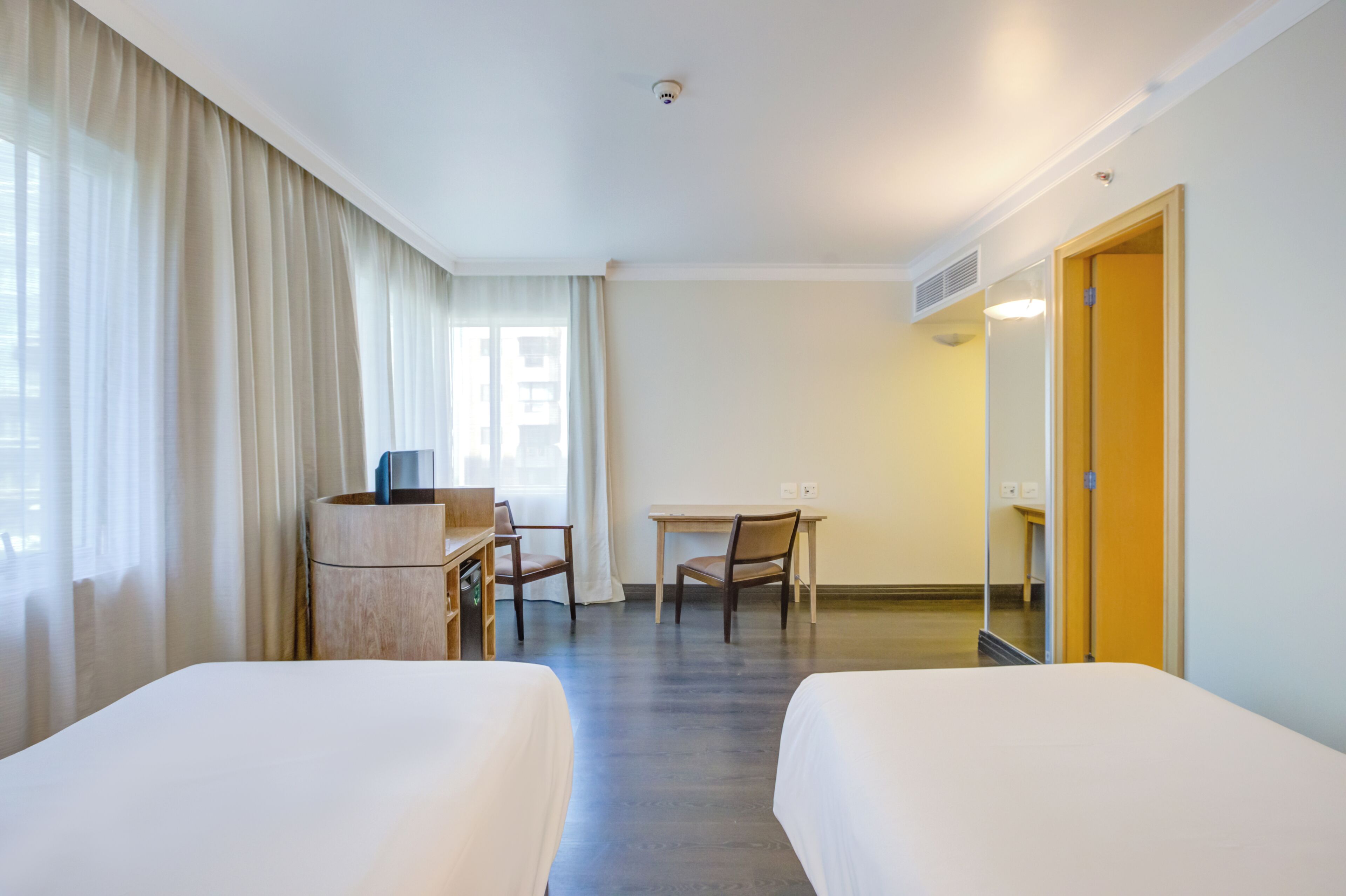 Quarto de hotel com duas camas, uma escrivaninha, uma cadeira, um pequeno armário de madeira e um espelho na parede.