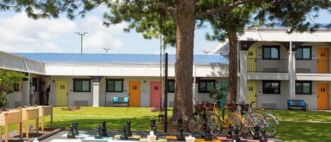 A large outdoor chessboard set up in front of a building with numbered doors, a tree, and bicycles parked nearby.