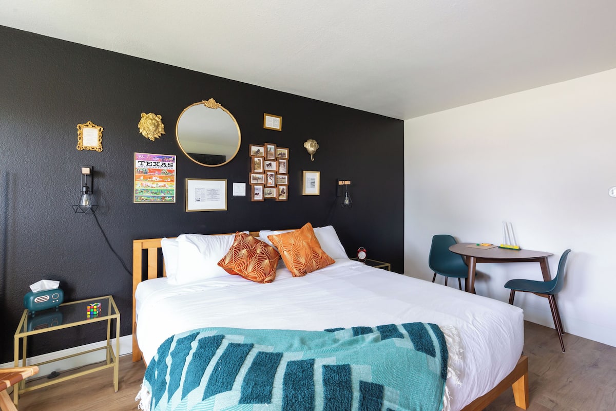 A neatly arranged hotel room with a large bed, a bedside table with a radio, a small stool, a round table with chairs, and a wall decorated with pictures and a mirror.