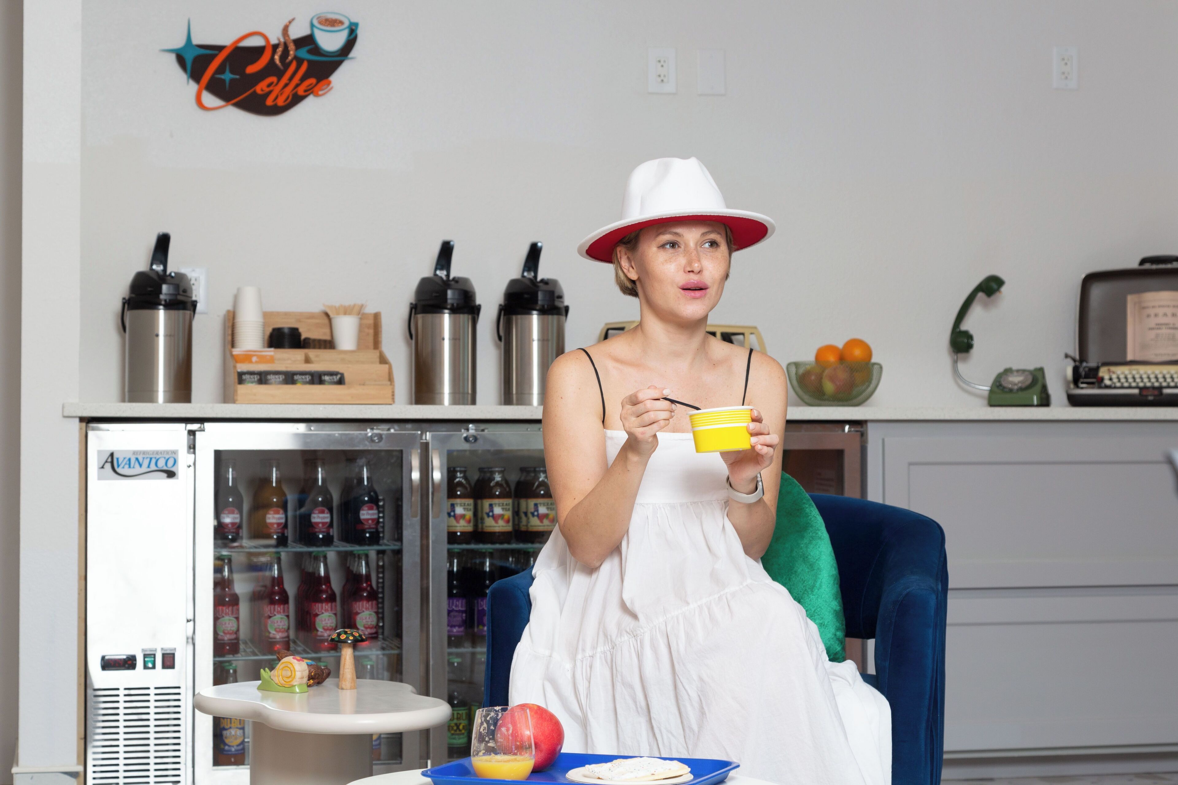 Una persona sentada en una habitación con un refrigerador lleno de bebidas, una mesita con un frutero y una bandeja de comida, y un teléfono antiguo sobre un escritorio.