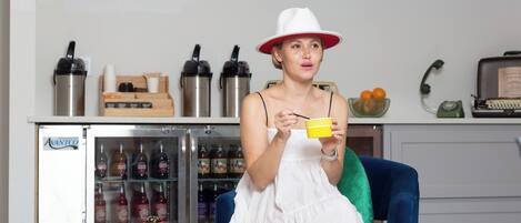 Una persona sentada en una habitación con un refrigerador lleno de bebidas, una mesita con un frutero y una bandeja de comida, y un teléfono antiguo sobre un escritorio.