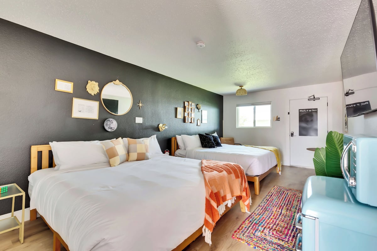 A hotel room with two beds, a mirror, wall decorations, a small table, and a suitcase.