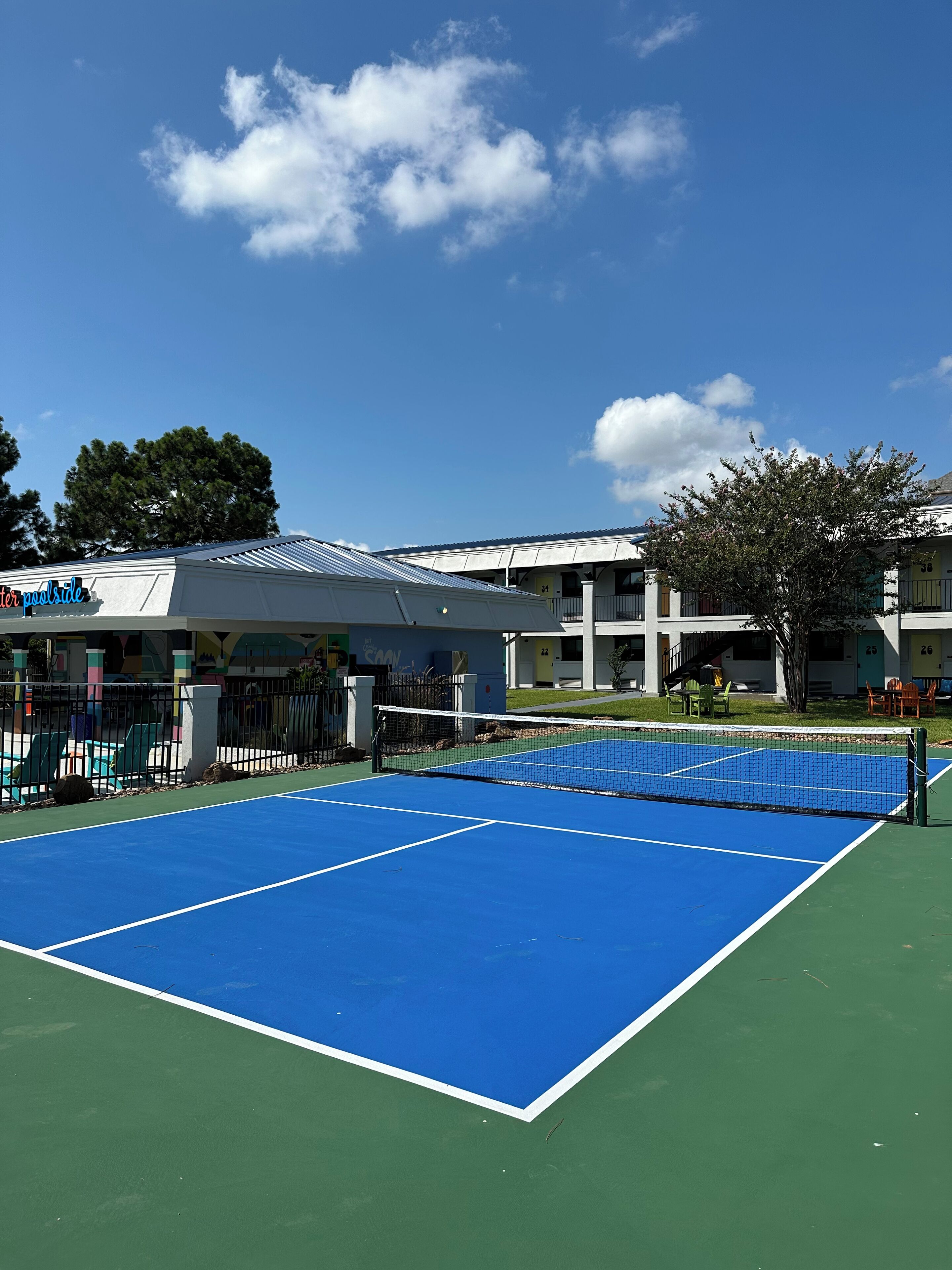 Een tennisbaan met een net, omringd door een hek, naast een gebouw met een bord waarop 'poolside' staat.