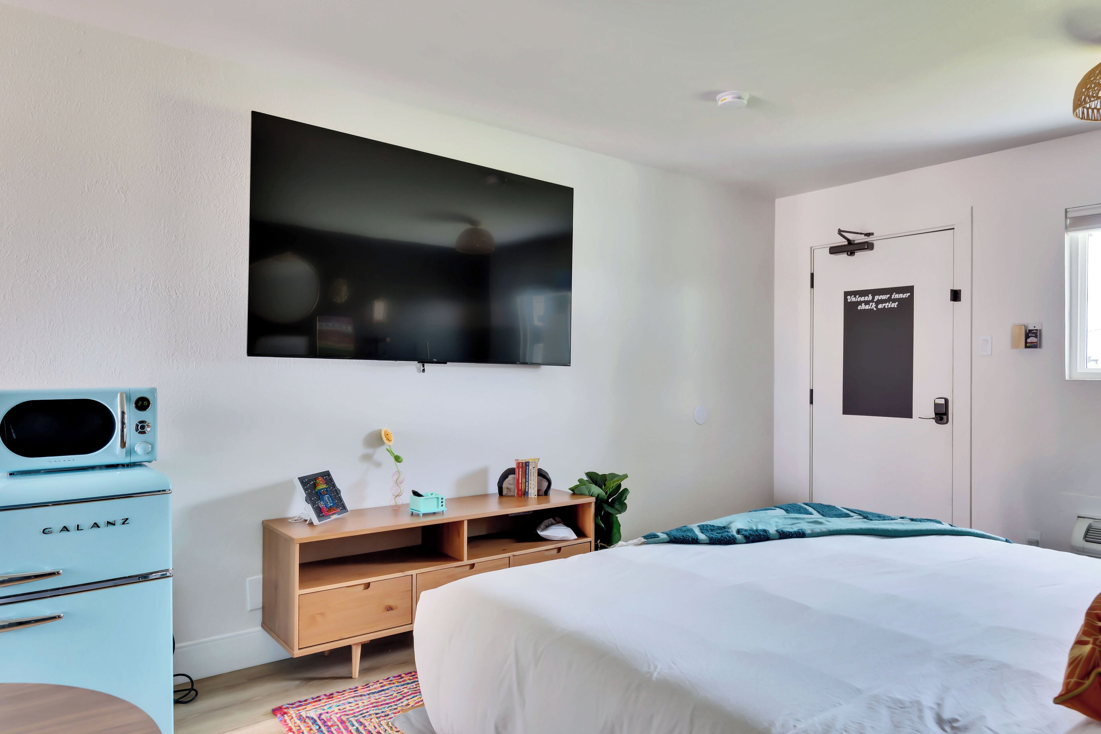 A bedroom with a bed, a television mounted on the wall, a small kitchenette with a blue appliance, a wooden dresser, and a door with a sign.