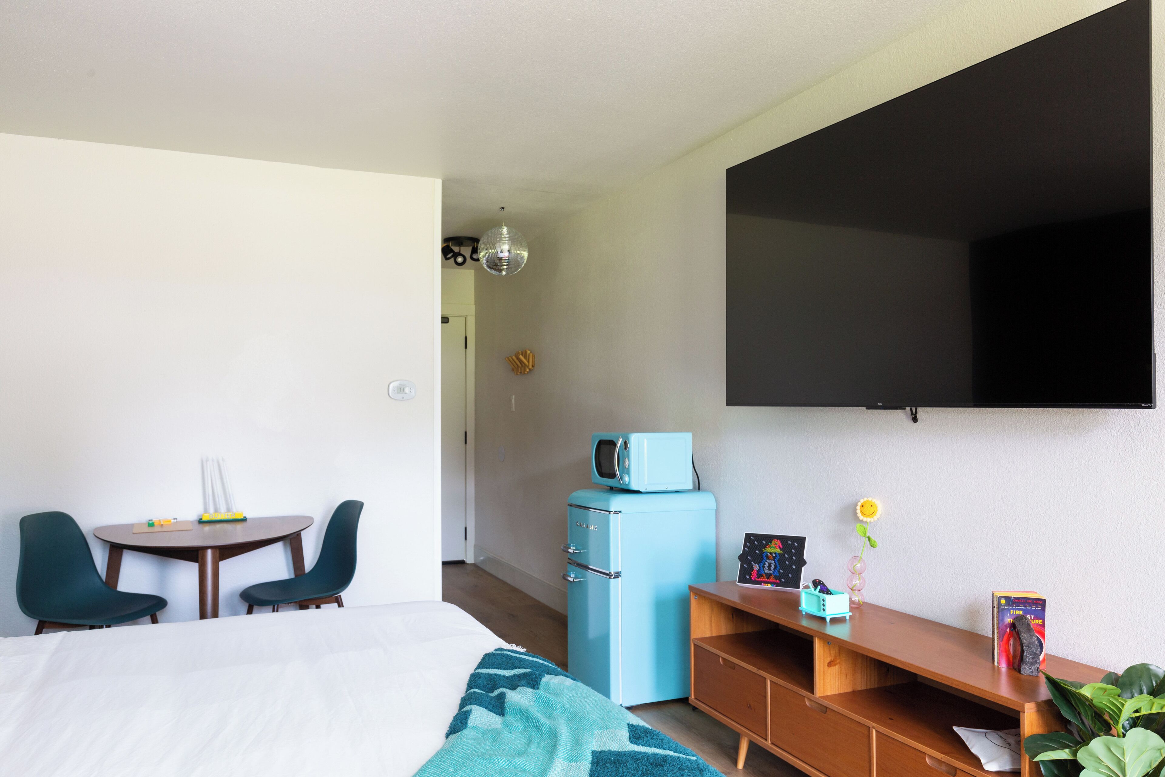 Habitación de hotel con cama, escritorio con silla, refrigerador pequeño, televisor montado en la pared y planta en maceta.