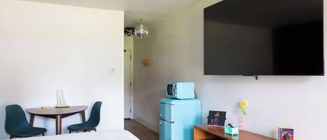 Habitación de hotel con cama, escritorio con silla, refrigerador pequeño, televisor montado en la pared y planta en maceta.