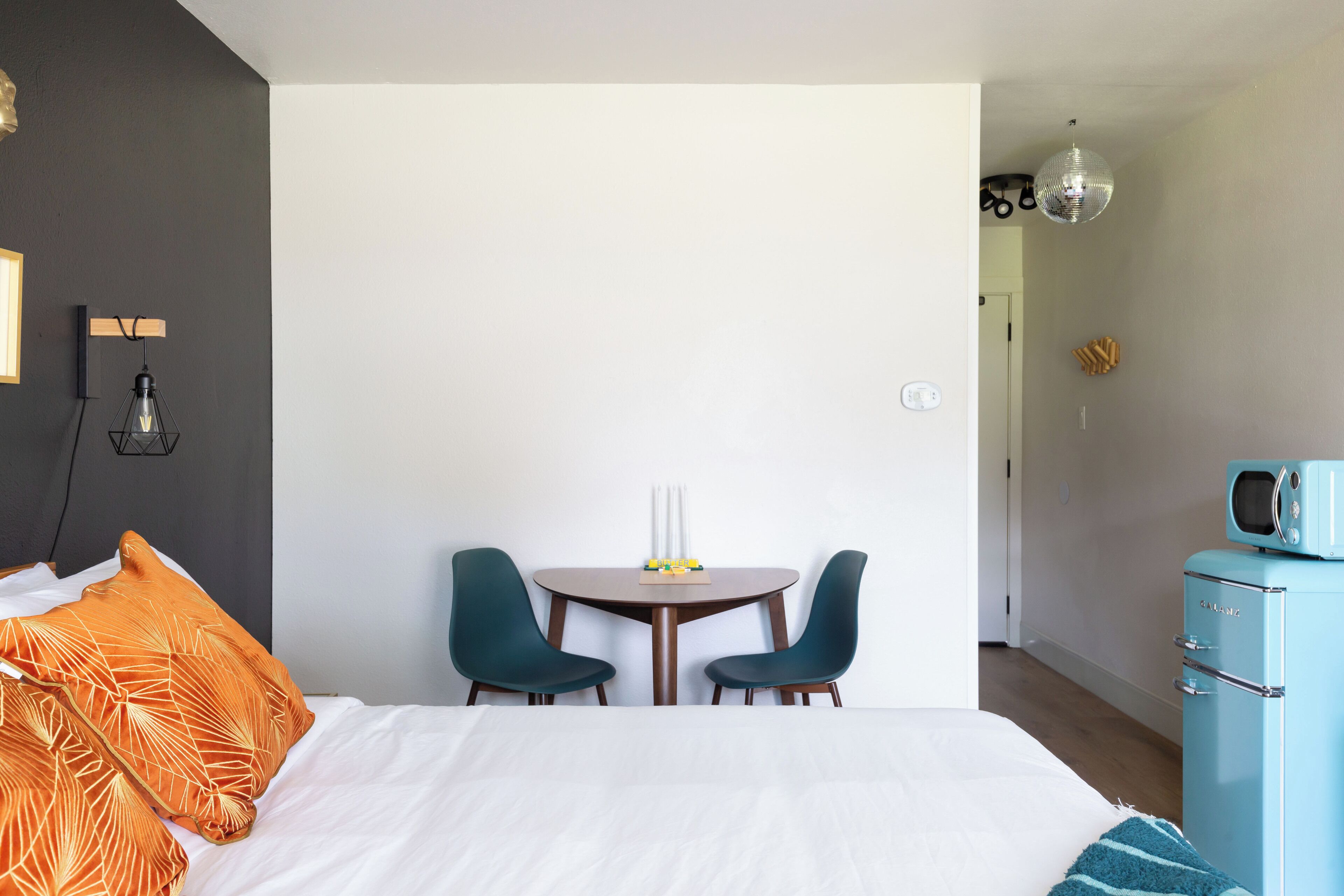 A modern hotel room with a large bed, two chairs, a small table, a vintage-style microwave, and a unique wall-mounted light fixture.
