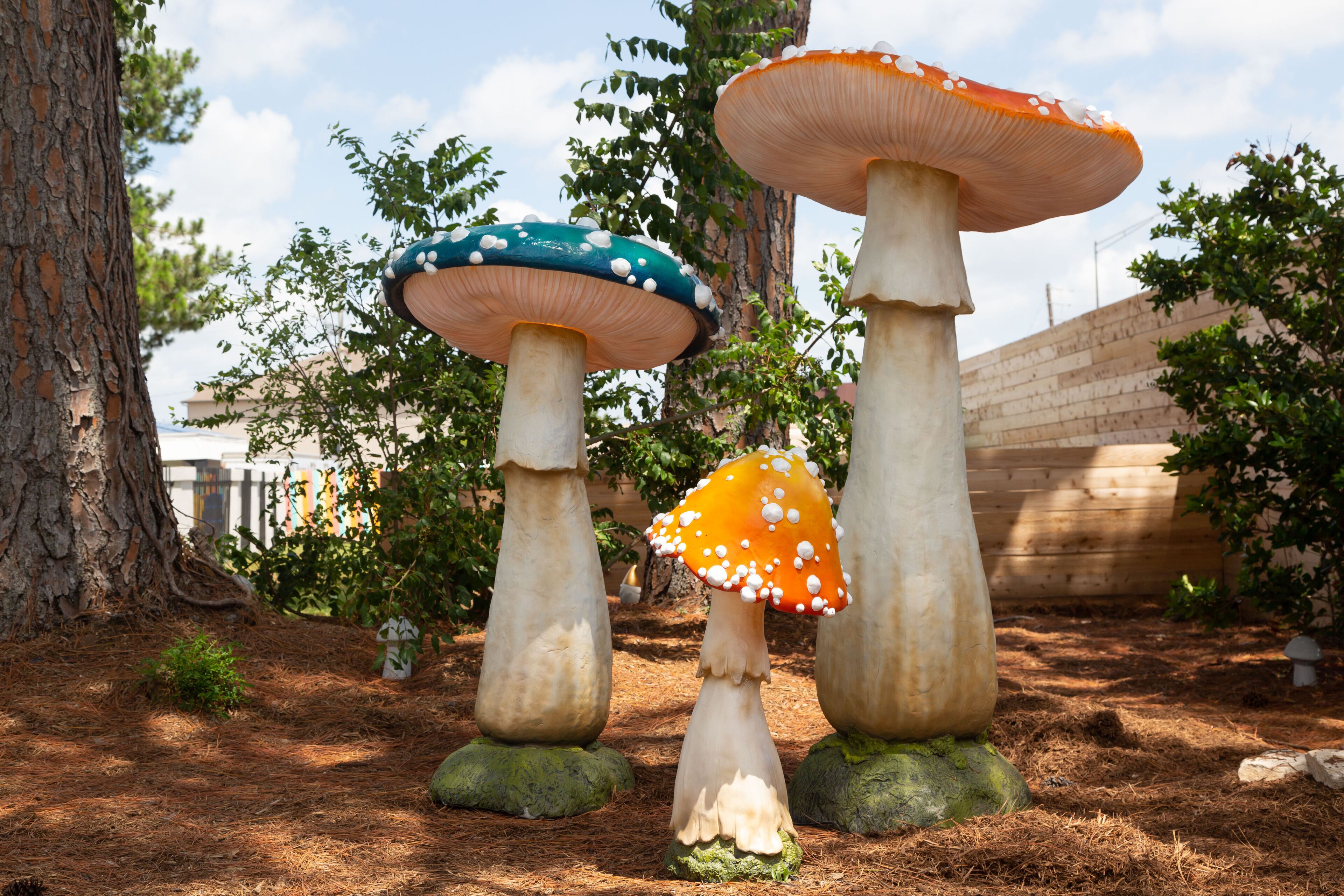 Three mushroom-shaped sculptures in a garden setting with trees and a brick wall in the background.