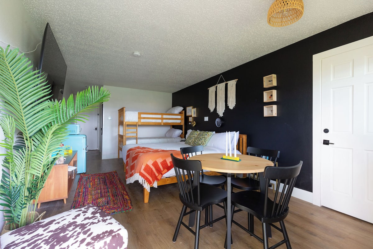 A compact room with a bunk bed, a dining table with chairs, a small kitchenette, and a large plant.