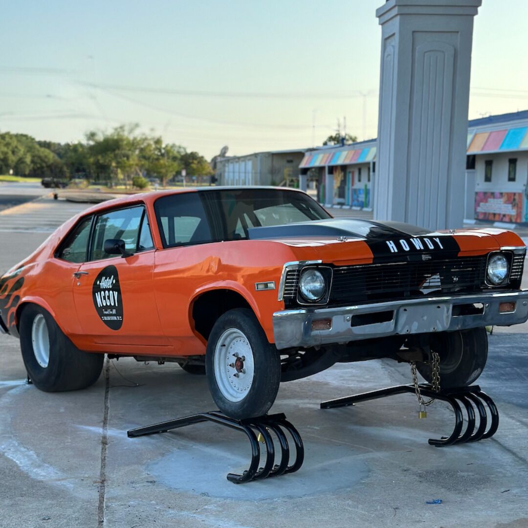 An orange vintage car with "HOWDY" and "Hotel NGCOV" decals parked on a concrete surface.