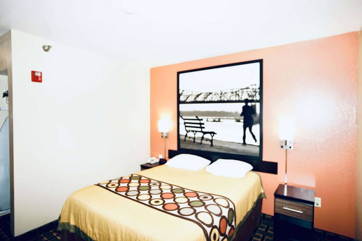 Habitación de hotel con cama, mesita de noche, lámpara y un cuadro en la pared que muestra un parque.