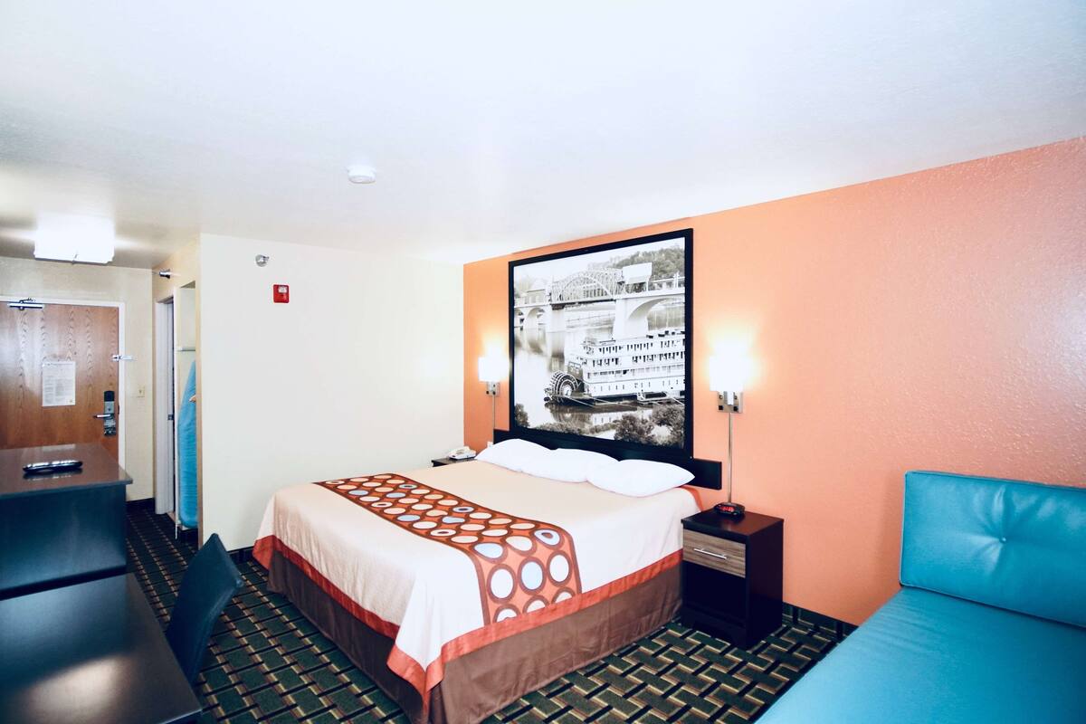 Habitación de hotel con cama, escritorio, silla, mesita de noche, lámpara y un cuadro colgado en la pared.
