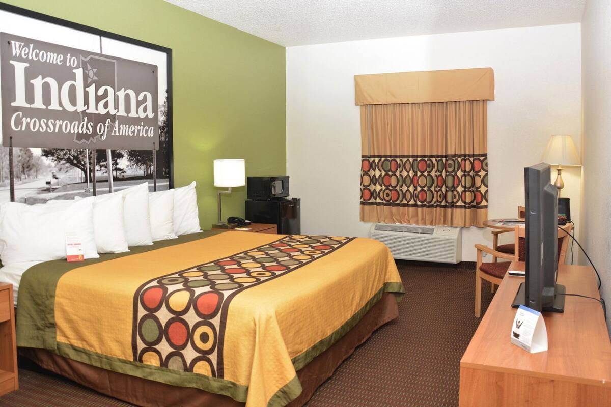 A hotel room with a bed, a desk with a computer, a TV, a window with curtains, and a sign reading 'Welcome to Indiana Crossroads of America.'