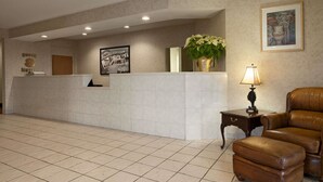 Uma área de recepção com piso de azulejos, balcão de atendimento, quadro decorativo na parede, planta em vaso, luminária e poltrona de couro.