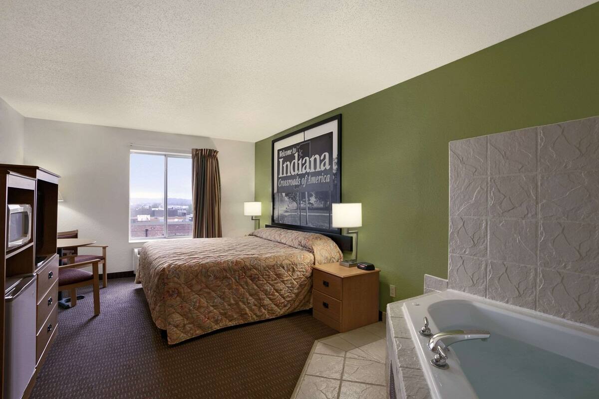 A hotel room with a bed, a desk, a chair, a window, a bathroom with a bathtub, and a framed welcome sign.