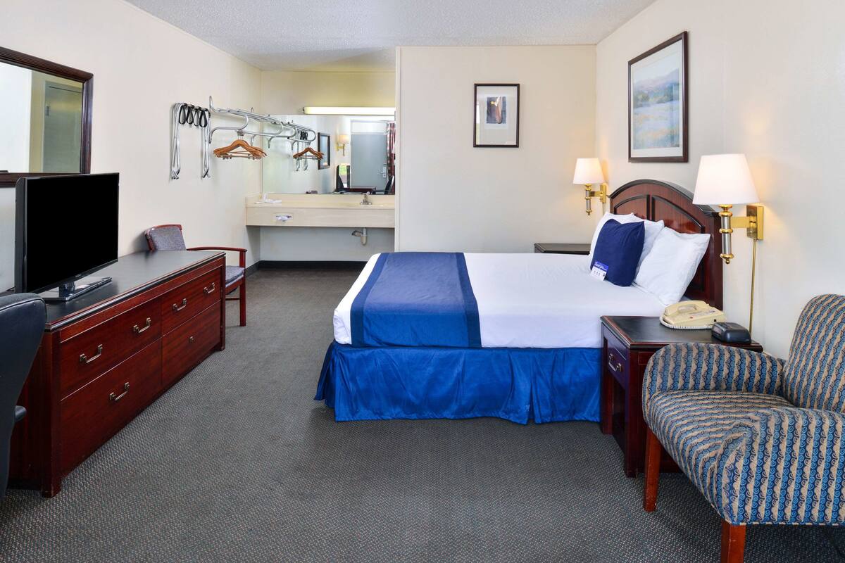 A hotel room with a bed, a desk with a television, a chair, a dresser, a mirror, and a bathroom with a sink and towel rack.