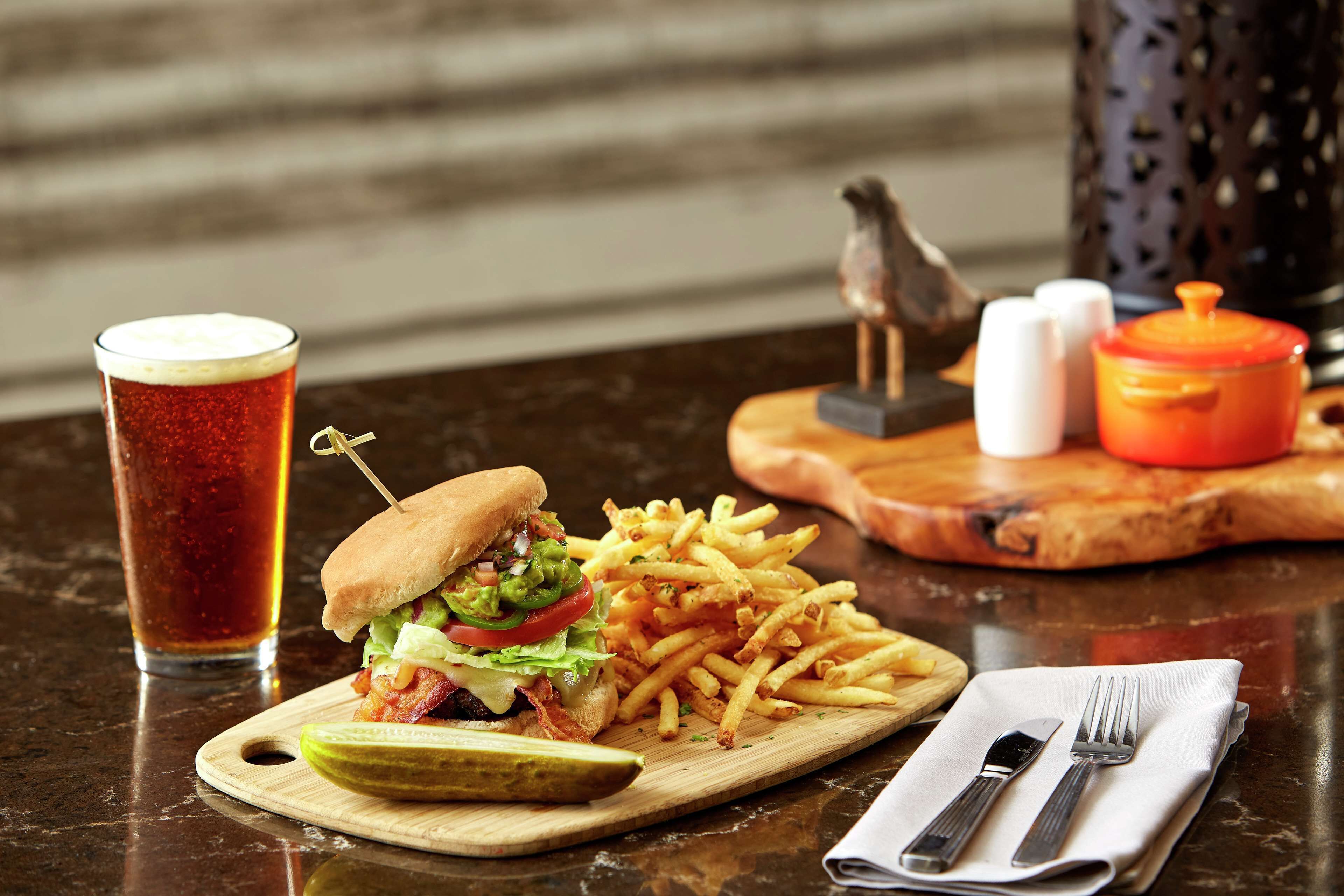 Un repas composé d’un hamburger, de frites et d’un cornichon, le tout servi sur une planche en bois, accompagné d’un verre de thé glacé et d’une serviette avec des couverts sur une table.