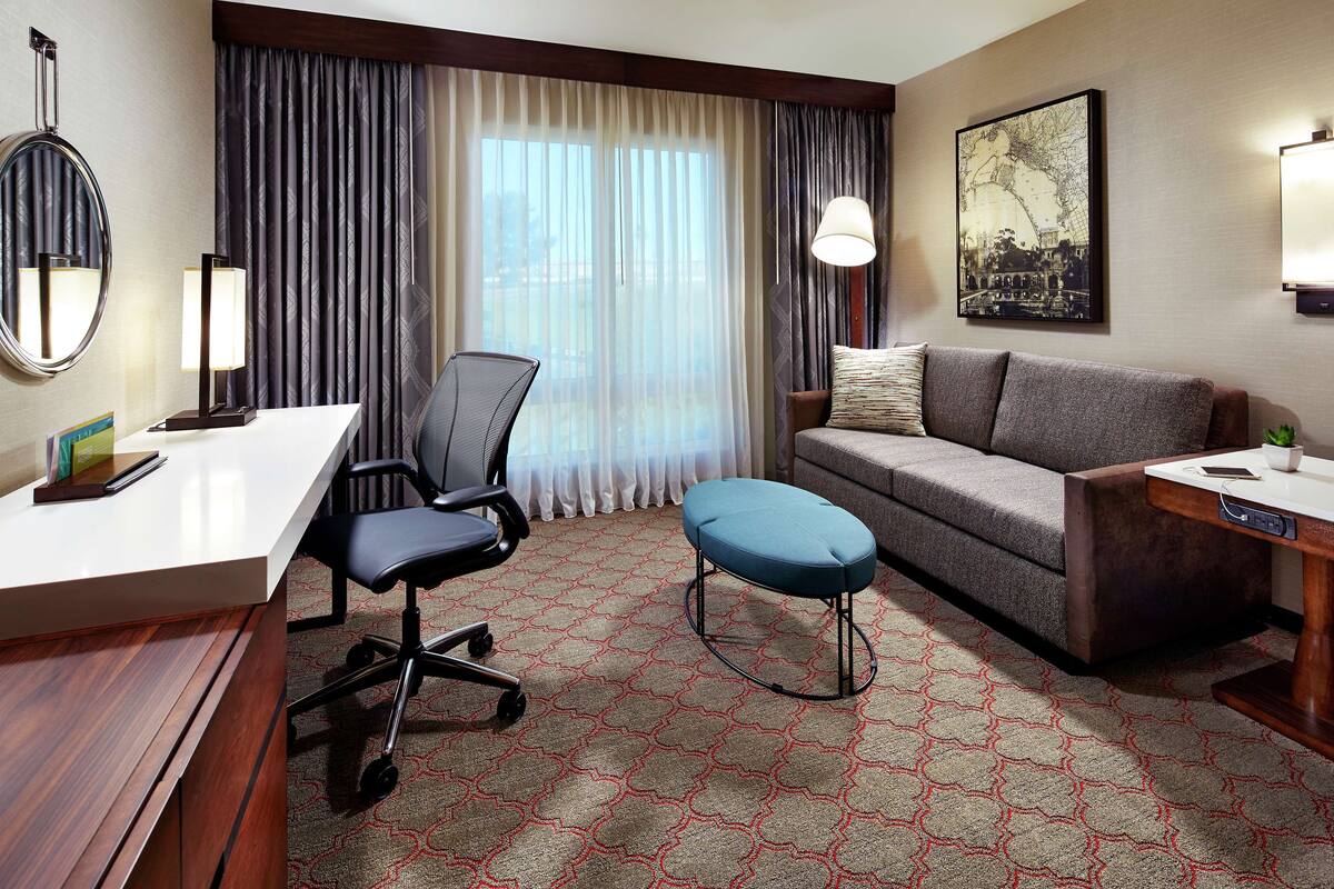 Habitación de hotel con escritorio, silla, sofá, reposapiés, lámpara, espejo y un ventanal grande con cortinas.