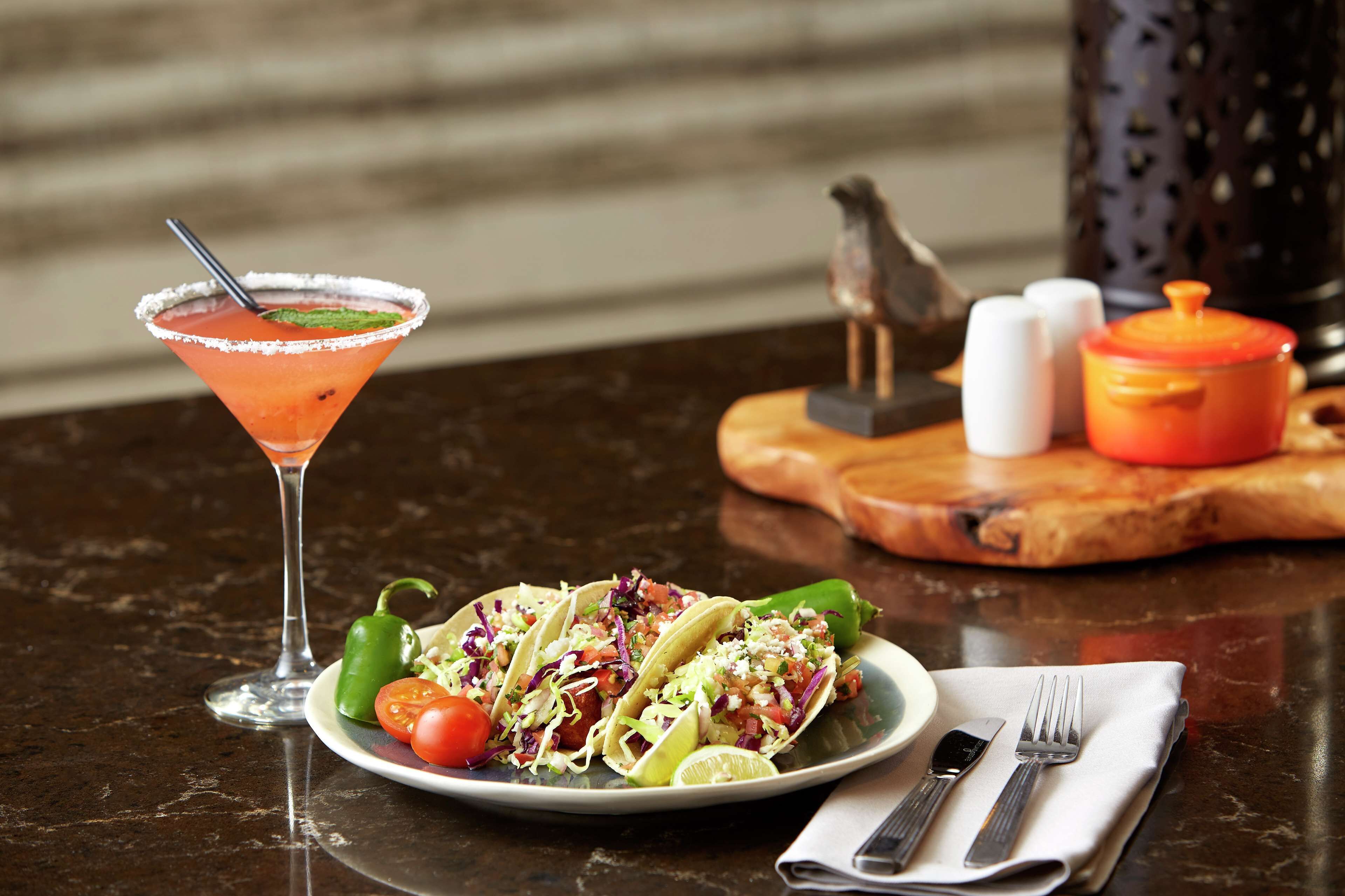 A plate of tacos with a lime wedge, a margarita glass, and a wooden cutting board with salt and pepper shakers.