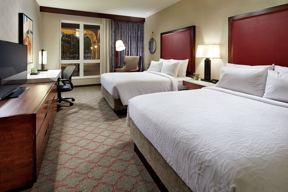 Ein Hotelzimmer mit zwei Betten, einem Schreibtisch mit Computer, einem Stuhl und einem großen Fenster mit Vorhängen.