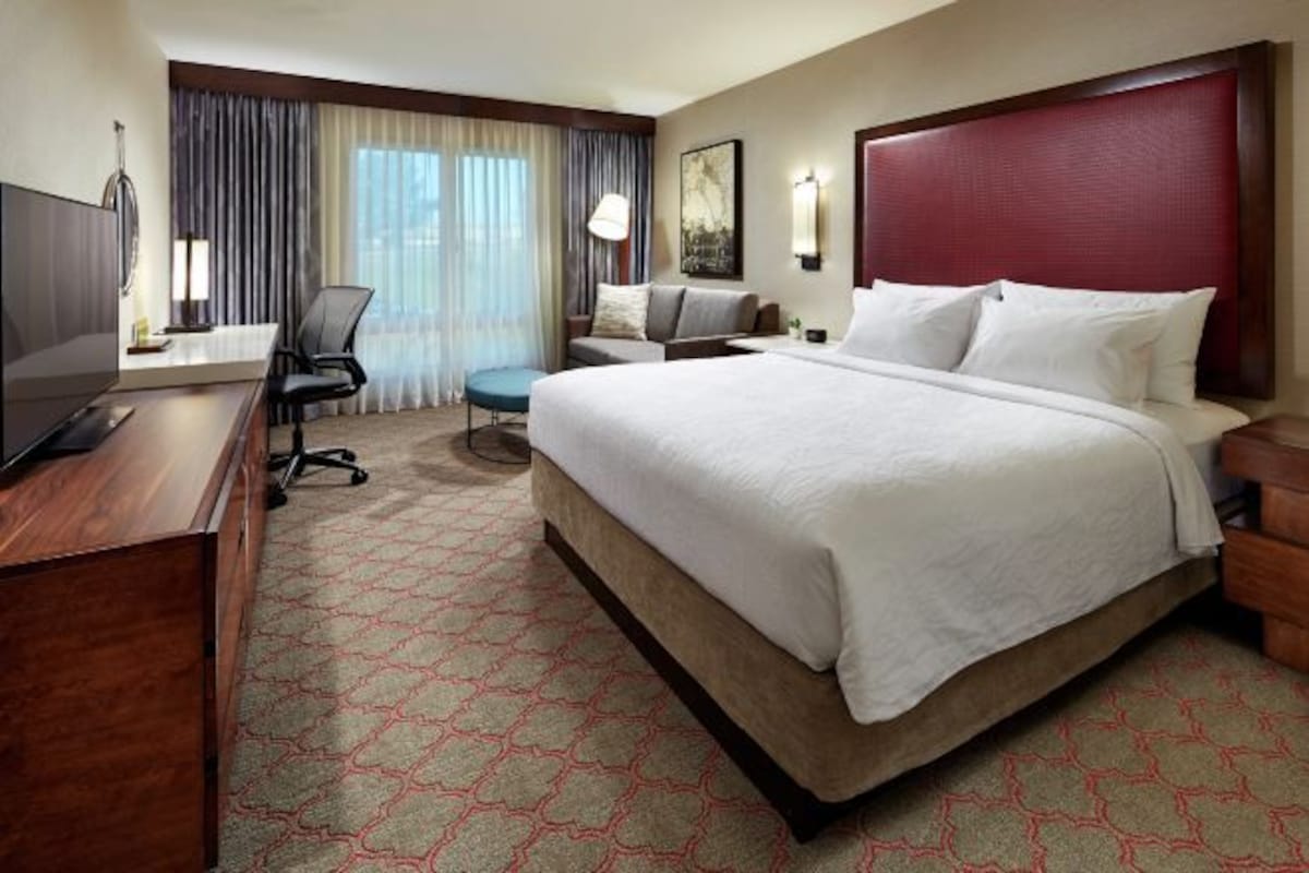 Une chambre d’hôtel avec un grand lit, un bureau avec un ordinateur, une chaise, une petite table et une fenêtre avec des rideaux.
