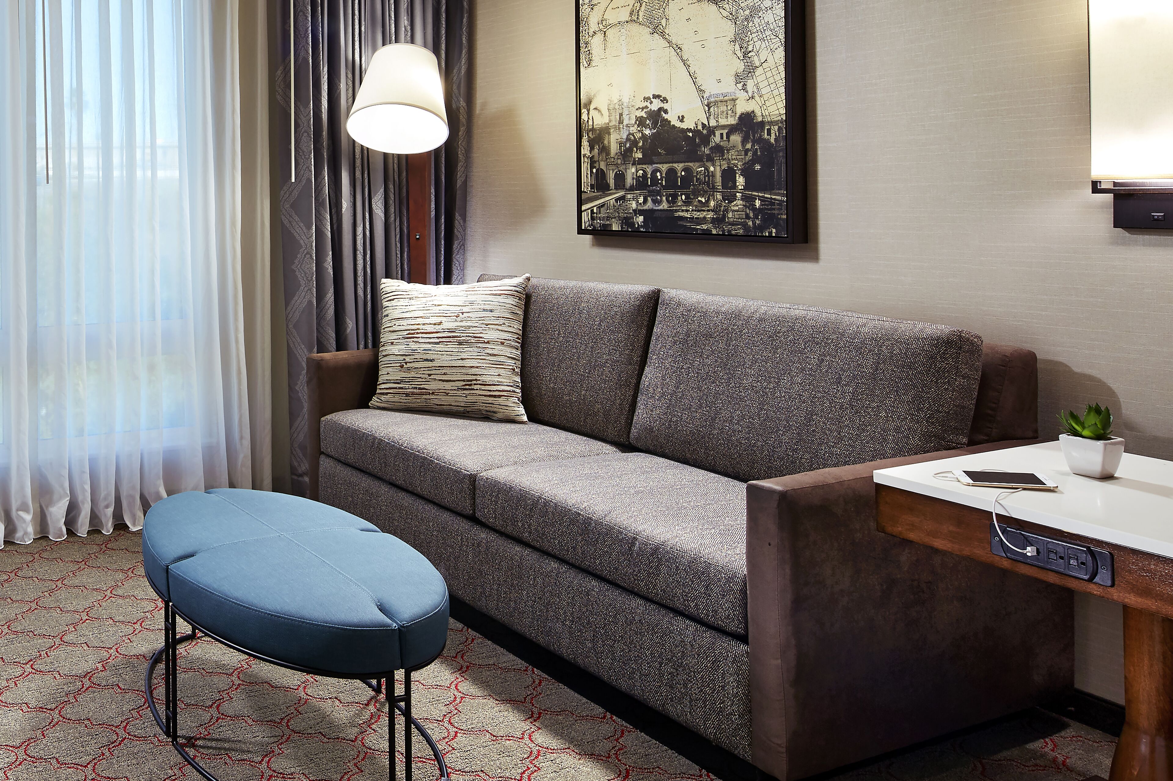 A modern hotel room with a grey sofa, a blue ottoman, a small table with a plant, and a wall-mounted lamp.