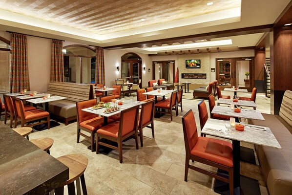 A spacious restaurant with multiple dining areas, each featuring tables and chairs. There are warm lighting fixtures on the ceiling, and a large mirror on one wall.
