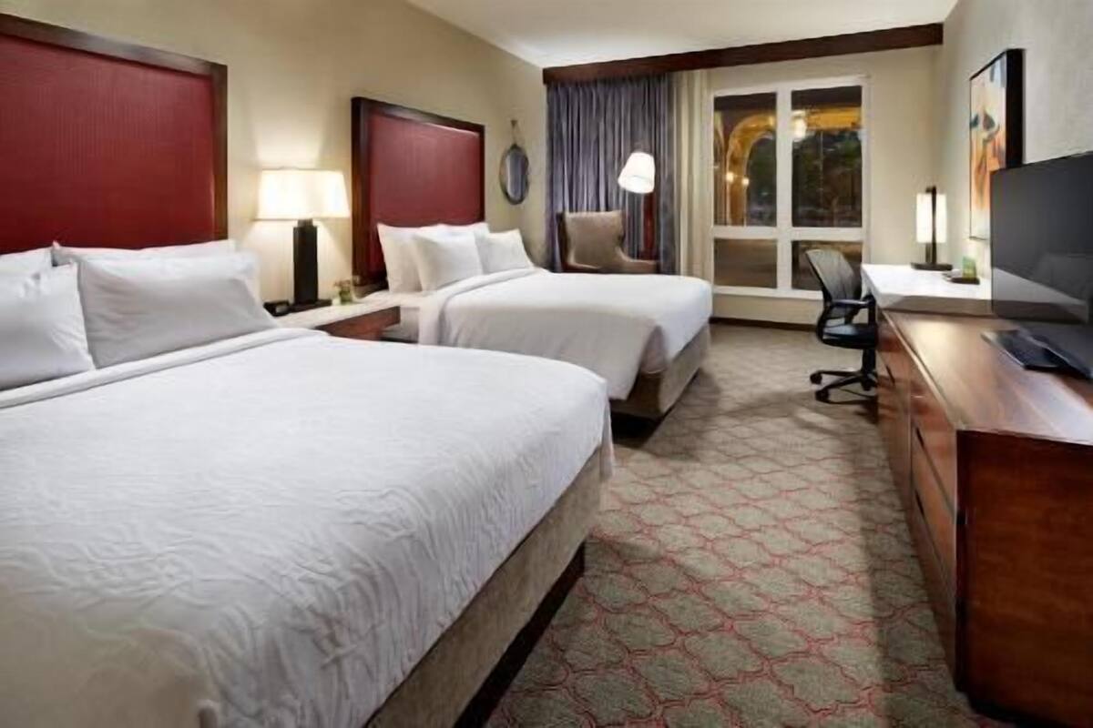 Ein Hotelzimmer mit zwei Betten, einem Schreibtisch, einem Stuhl, einem Fernseher und einem Fenster mit Vorhängen.