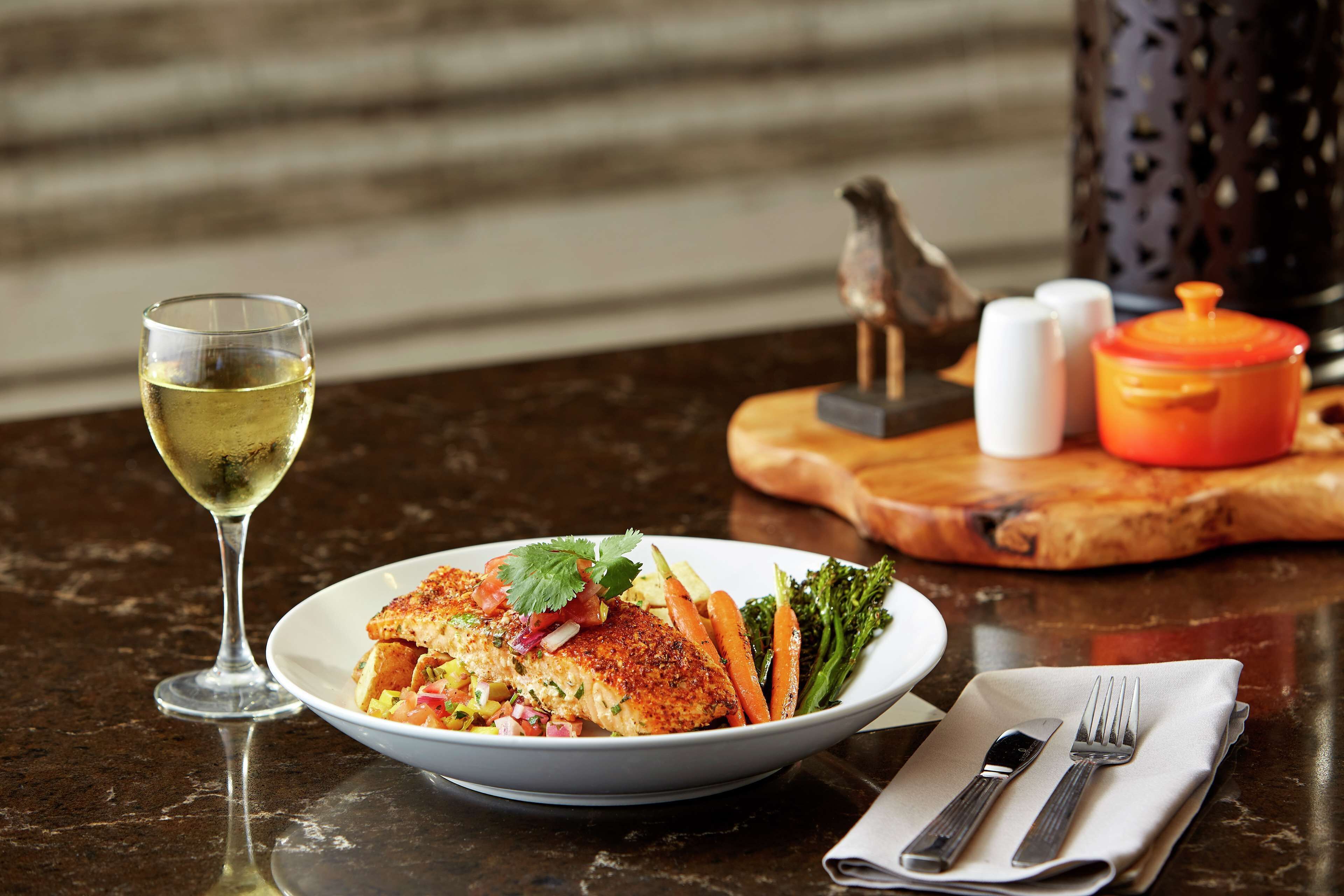 A plate of food with a glass of white wine, salt and pepper shakers, a small bird figurine, and a red pot on a wooden board.