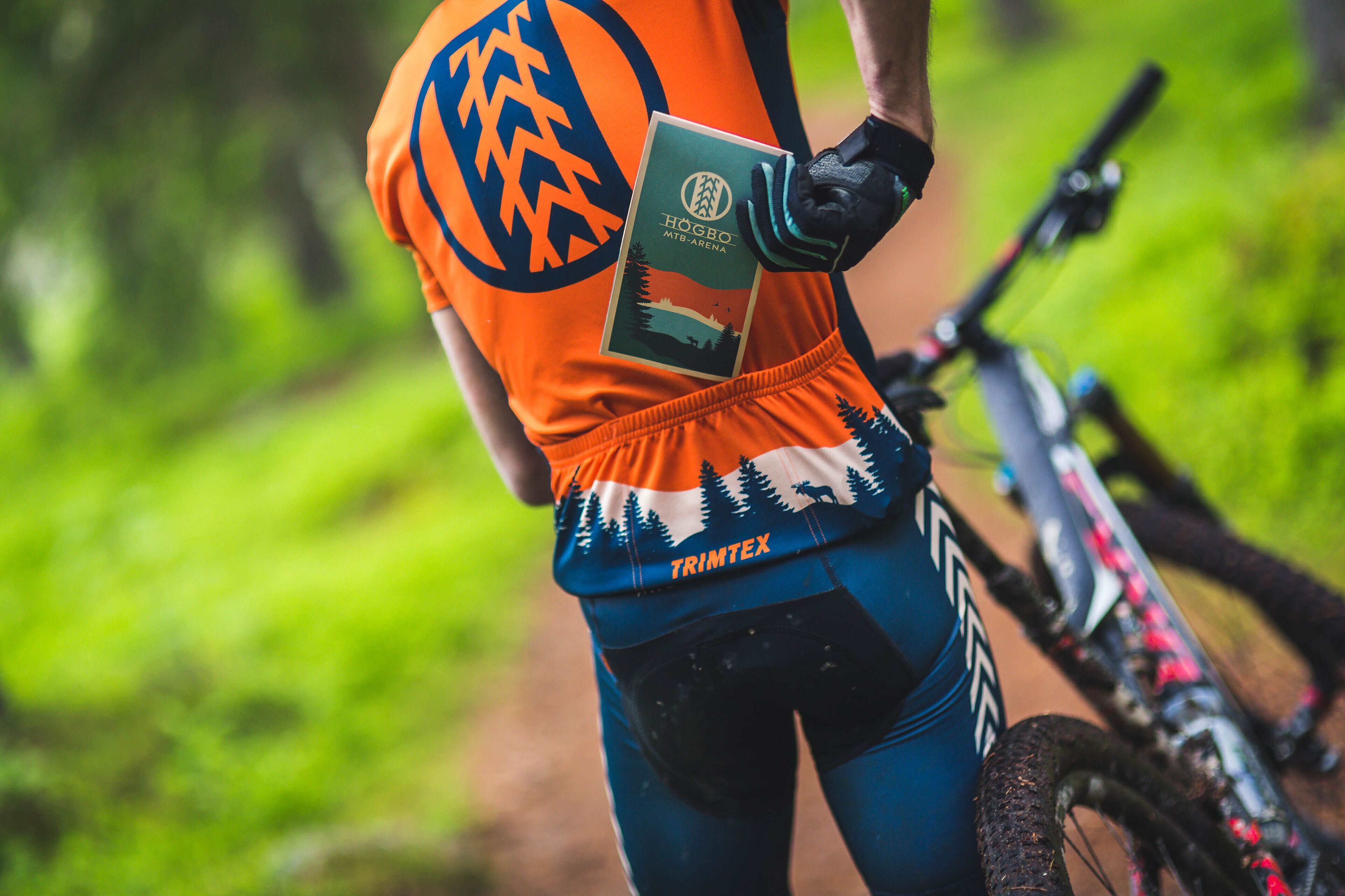 Een persoon in fietspak houdt een startnummer en een waterfles vast, met een mountainbike op de achtergrond.