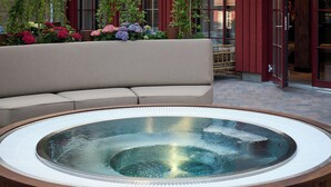 Un jacuzzi con estructura de madera y tapa de cristal, rodeado por un sofá beis y plantas en macetas.
