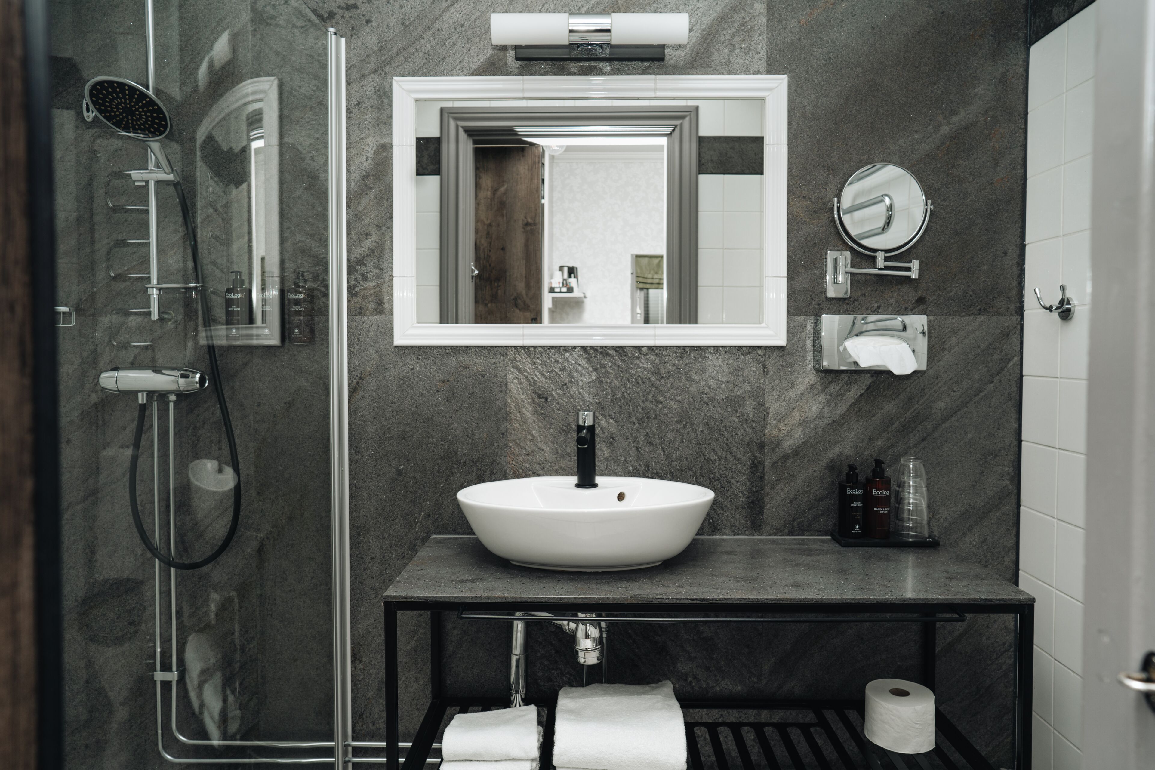 A modern bathroom with a large mirror, a vessel sink, a handheld showerhead, and a toilet paper holder.