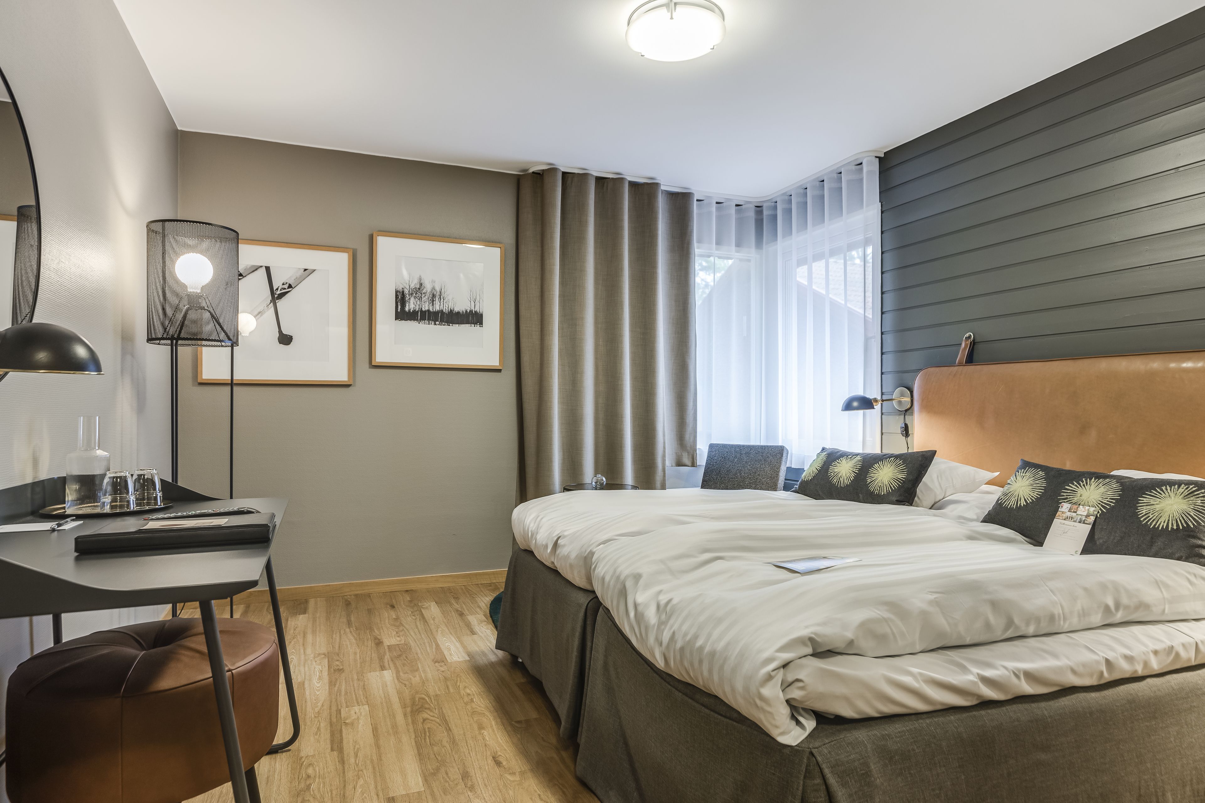 A modern hotel room with a bed, desk, and chair. There are framed pictures on the wall, a window with curtains, and a ceiling light.
